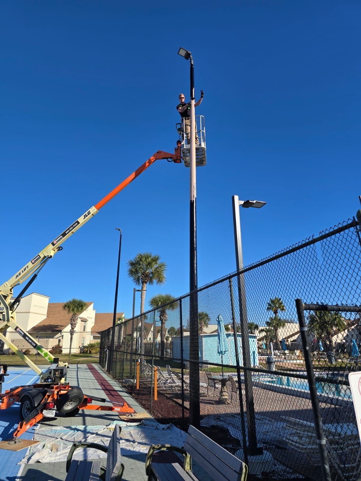 Person in cherry picker working on a black light pole by a pool with a fence and palm trees.