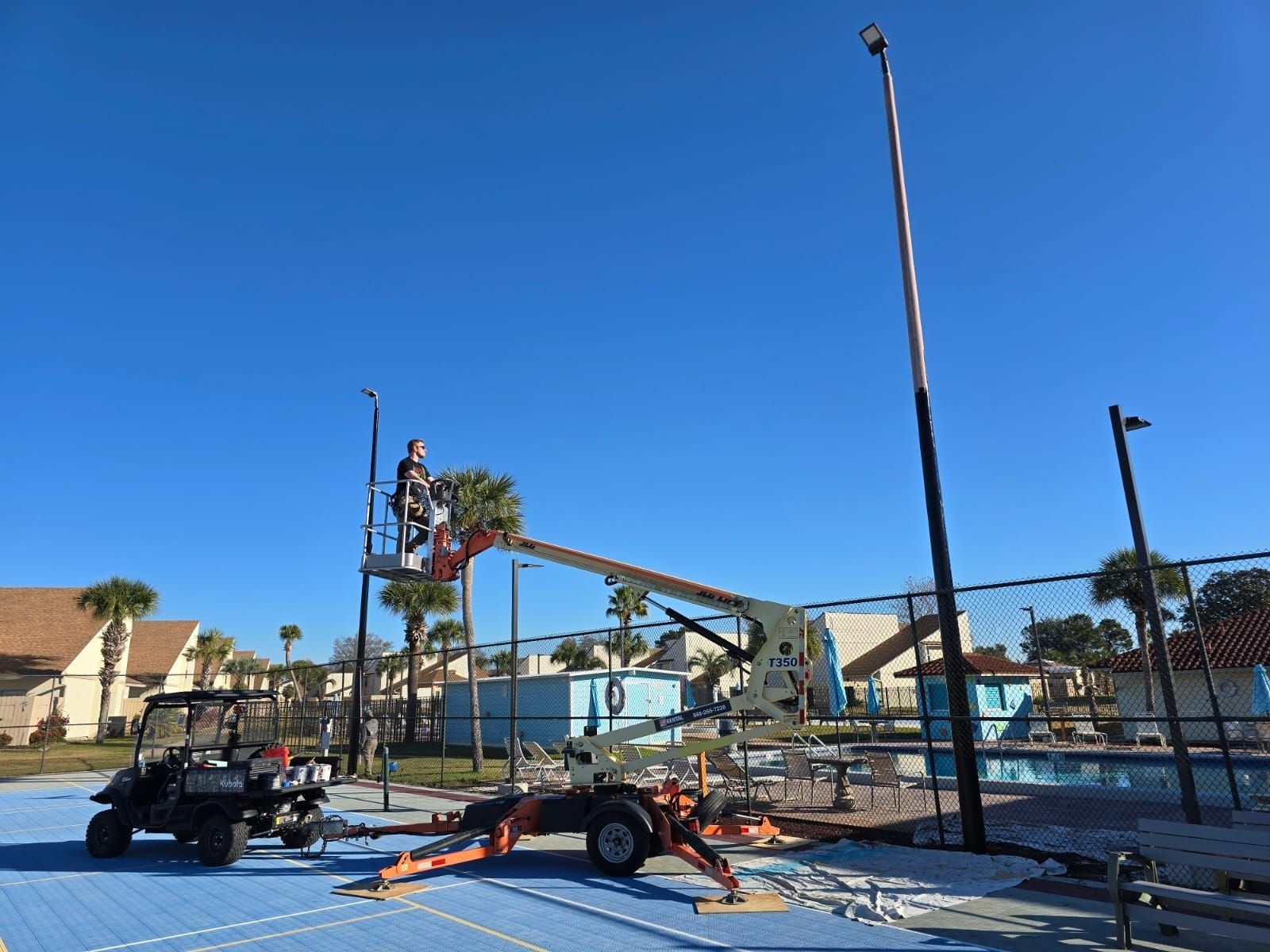 Workers in a lift installing a tall light pole near a pool, sunny day.
