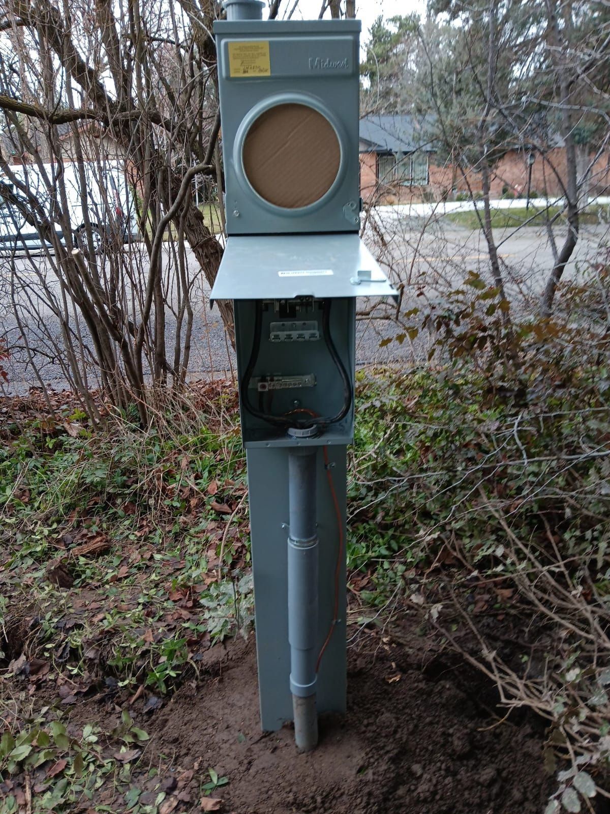 Outdoor utility box with a circular mesh top, metal gray, planted in brown earth, surrounded by green bushes.
