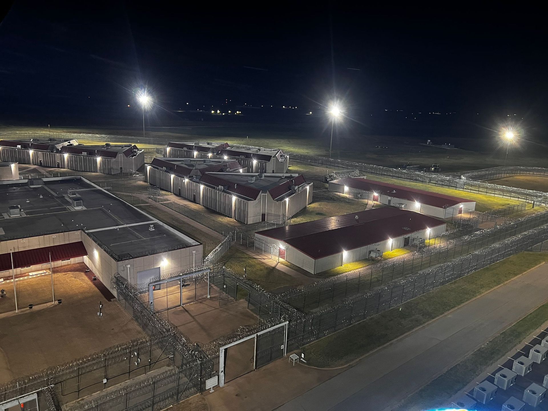 Nighttime aerial view of a prison complex with illuminated buildings, fences, and spotlights.
