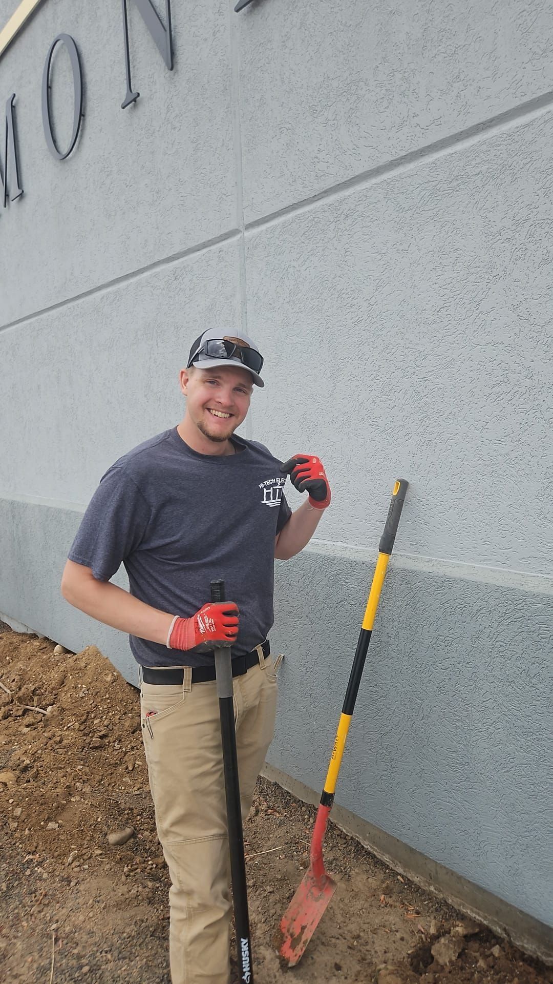 Man in work clothes smiles next to a wall, holding a shovel. He wears a hat and gloves in an outdoor setting.