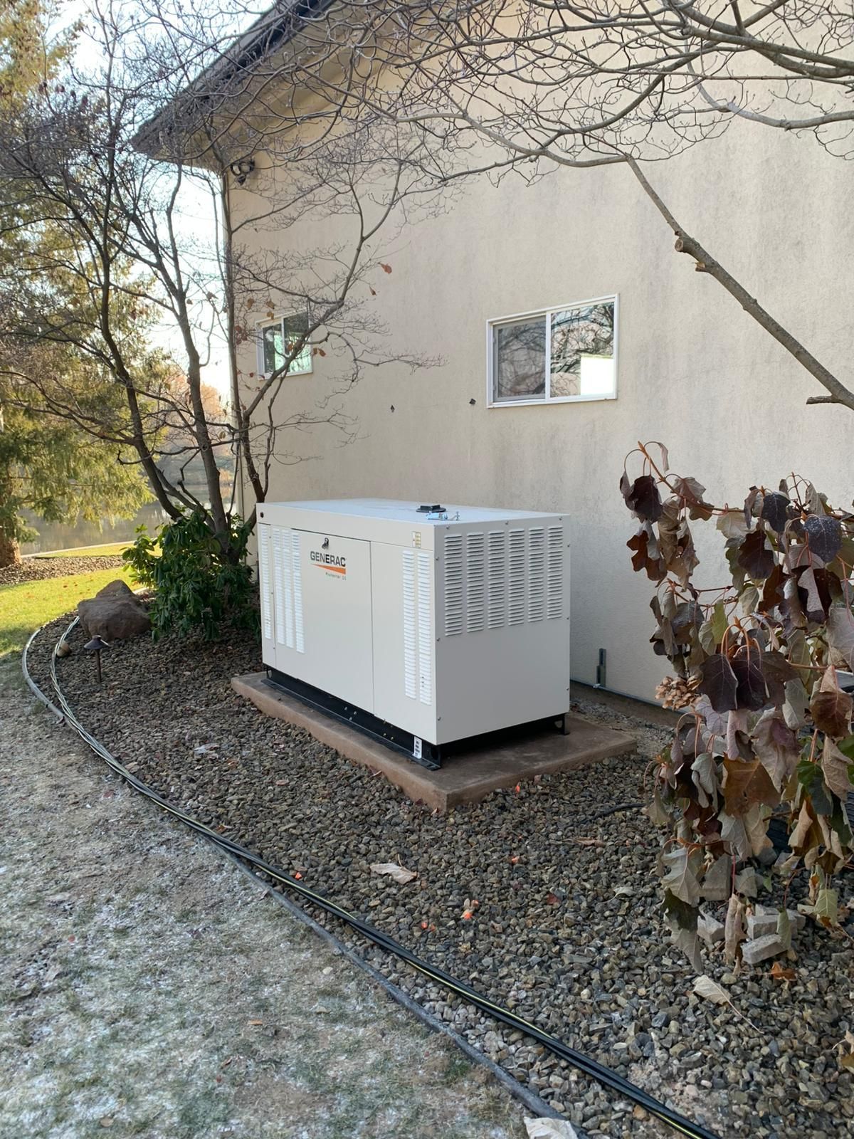 Generator unit, beige, on a concrete pad next to a light-colored house. Bare trees and gravel surround it.