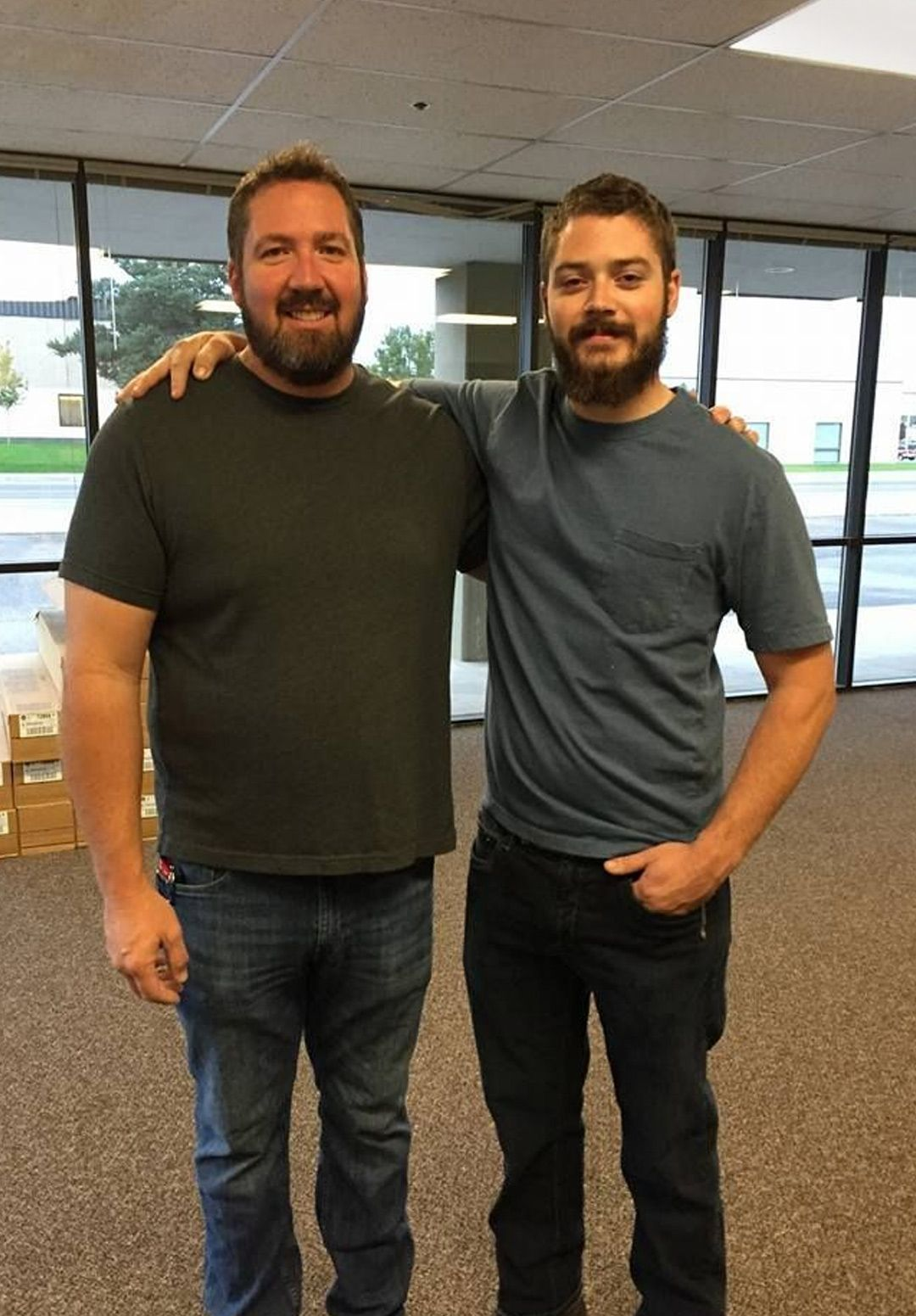 Two men with beards pose indoors, one arm over the other's shoulder, in front of windows.