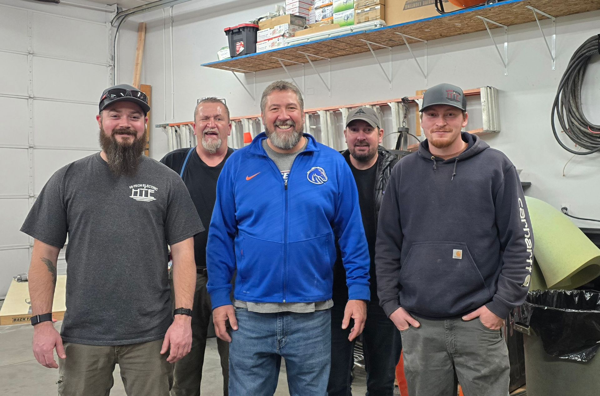 Five men standing together inside a workshop. Two are wearing baseball caps. One man smiles broadly, wearing a blue jacket.