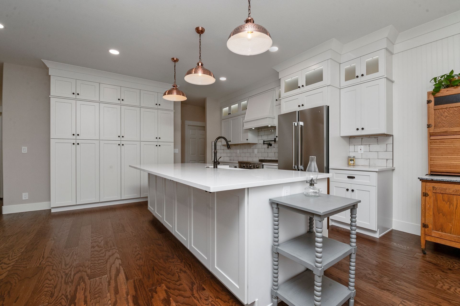 White kitchen with island, cabinets, copper lights, stainless steel appliances, and dark wood floors.