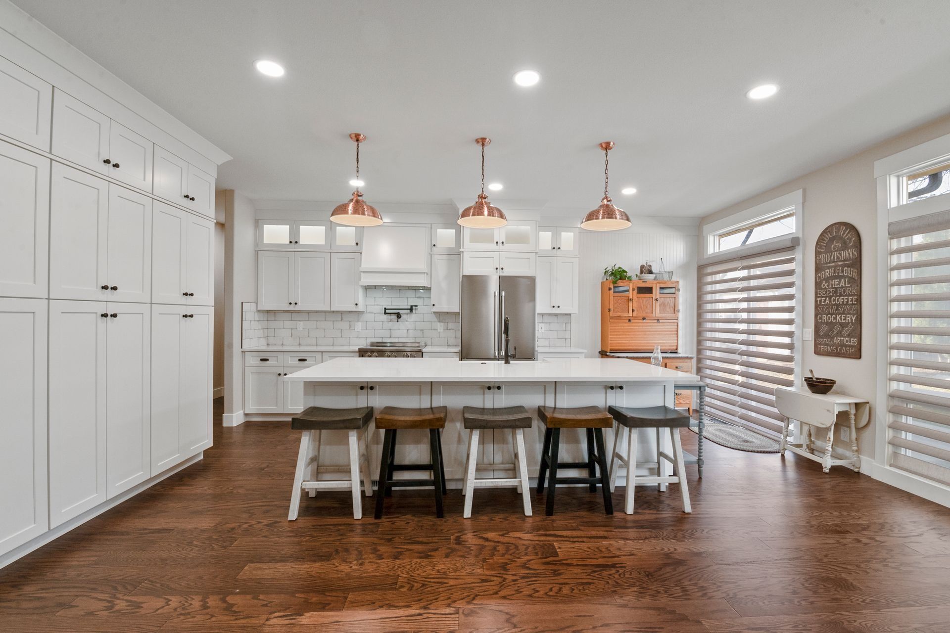 White kitchen with island, wood floor, copper lights, white cabinets and counters, and dark wood stools.