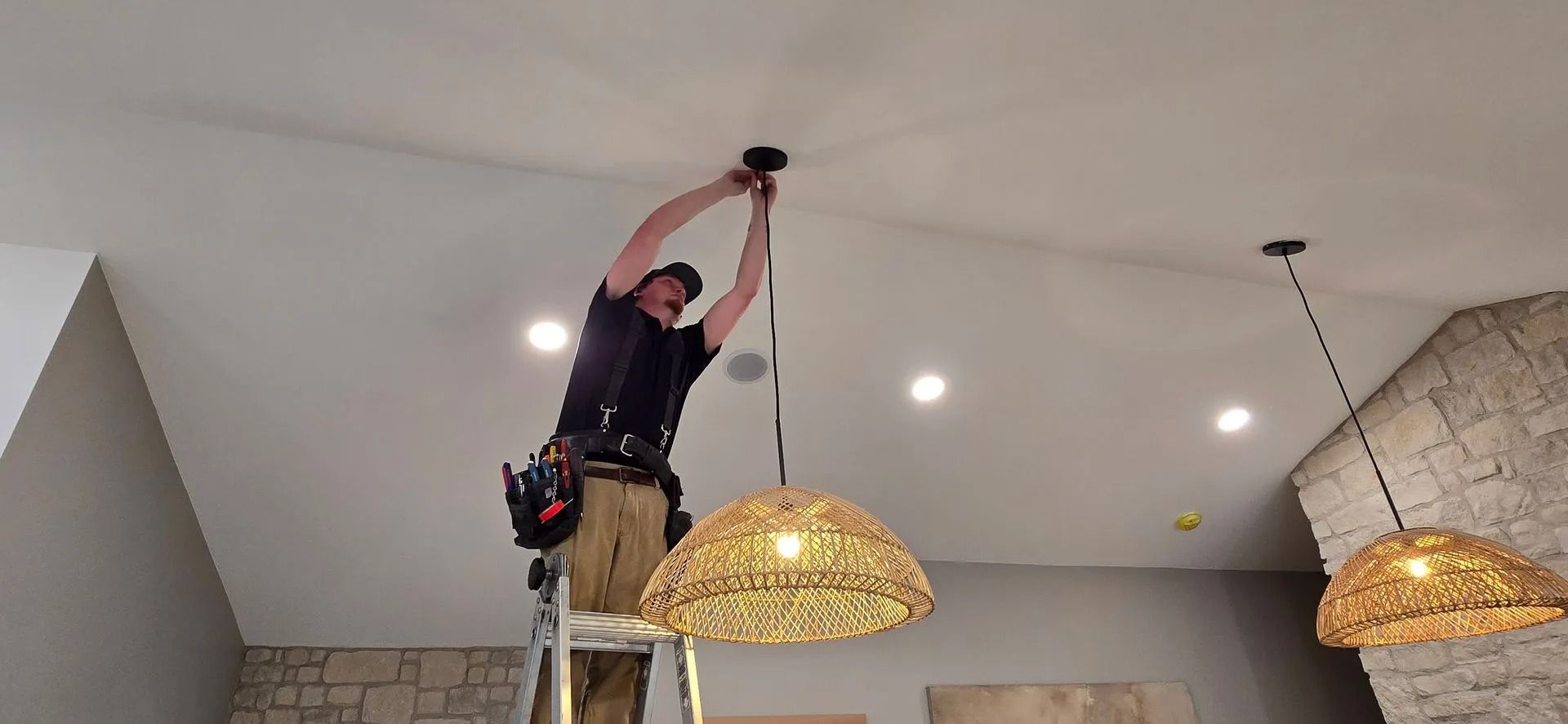 An electrician installing a pendant light, standing on a ladder. White ceiling, stone wall.