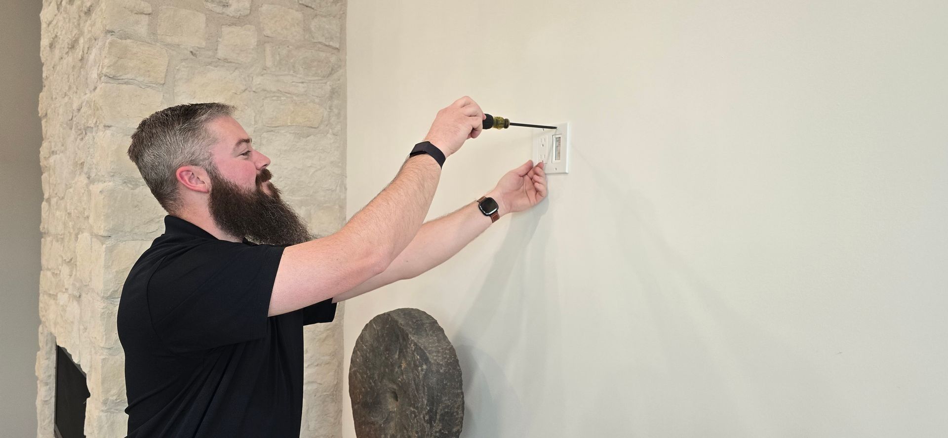 A man with a beard uses a screwdriver on an electrical outlet on a light-colored wall.