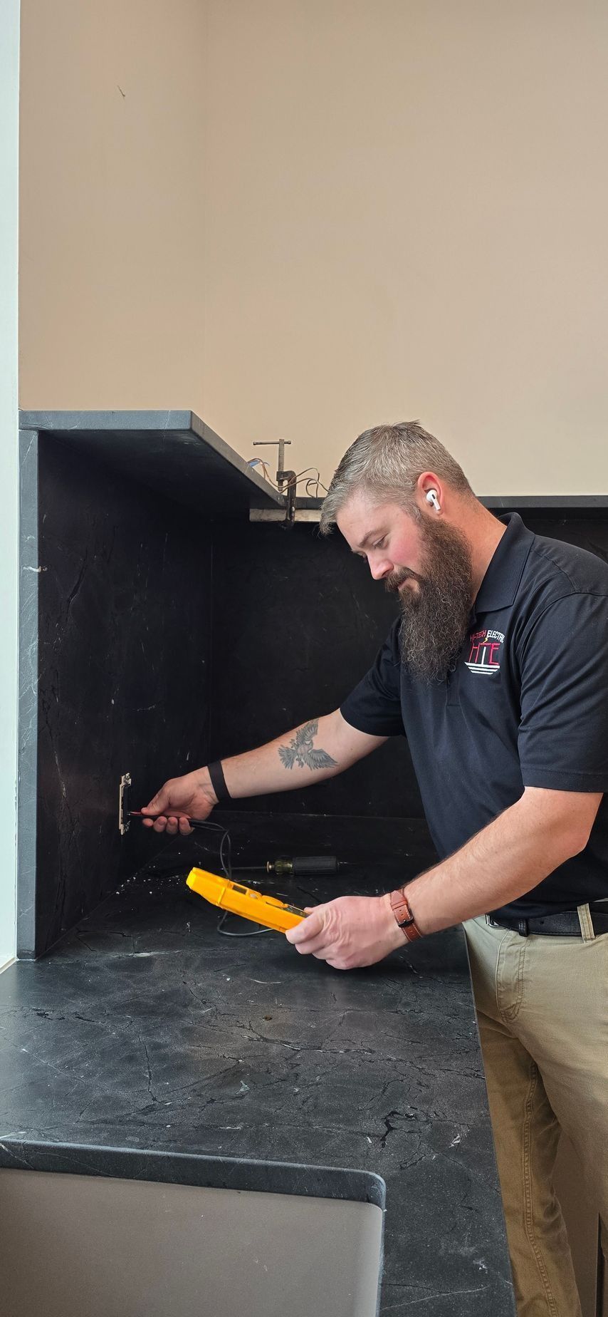 A man with a beard checks an outlet with a yellow voltage tester. He's in front of a black counter, near a tan wall.