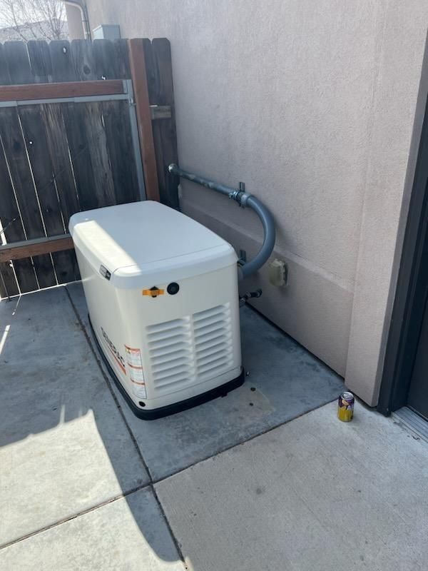 White standby generator next to a beige building and a wooden fence, on a concrete pad.