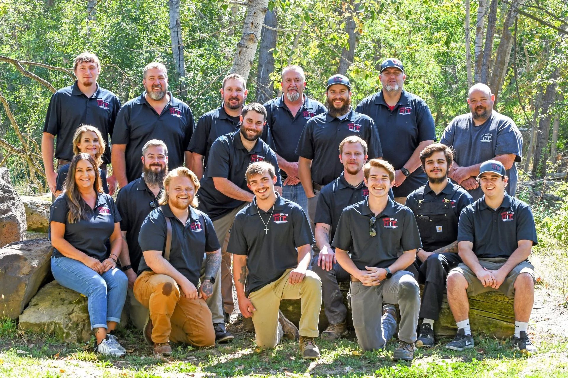 Group of people wearing black shirts, posing outside in front of trees.