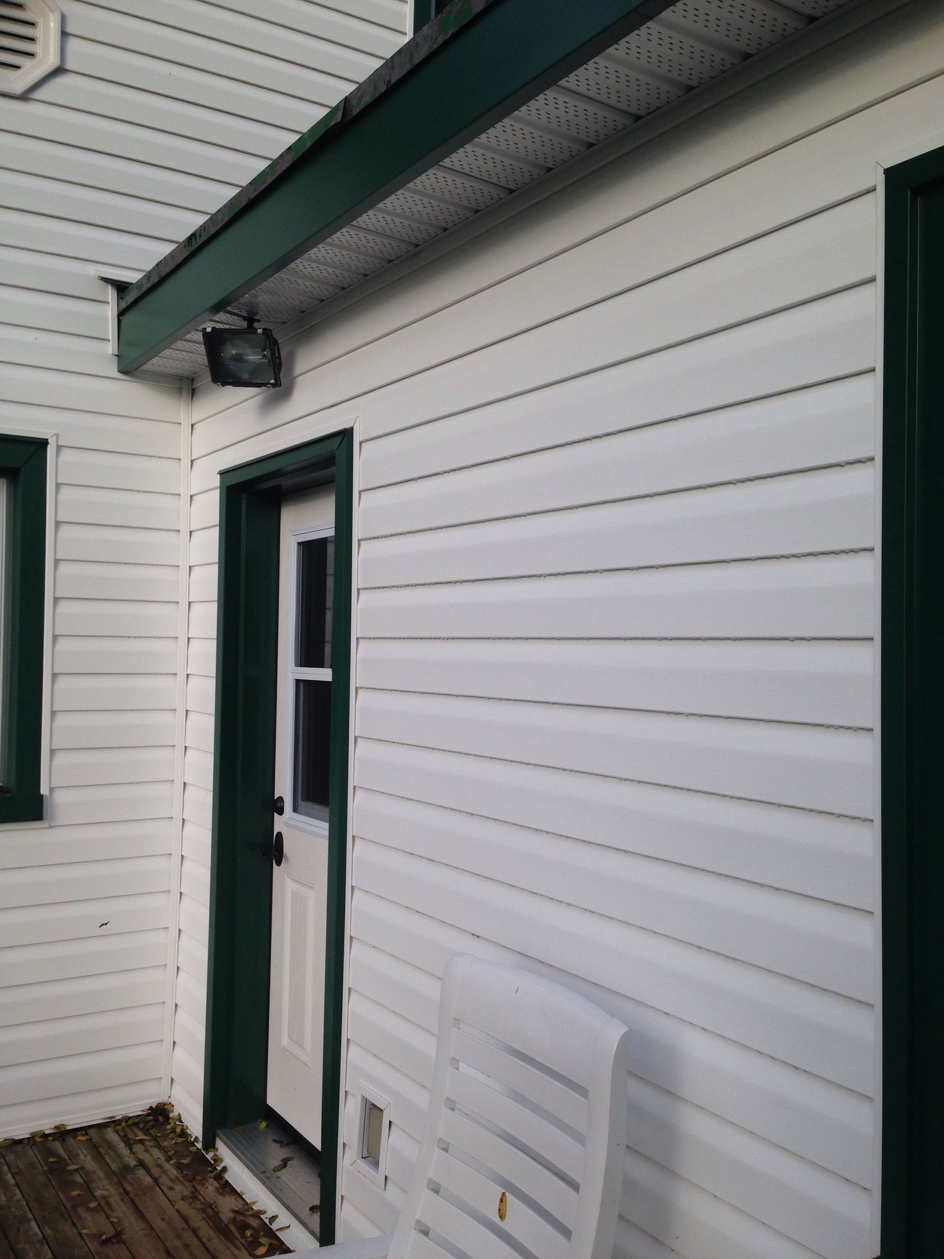A white-sided building exterior with a green-trimmed door, a motion light above, and a white plastic chair on the deck.