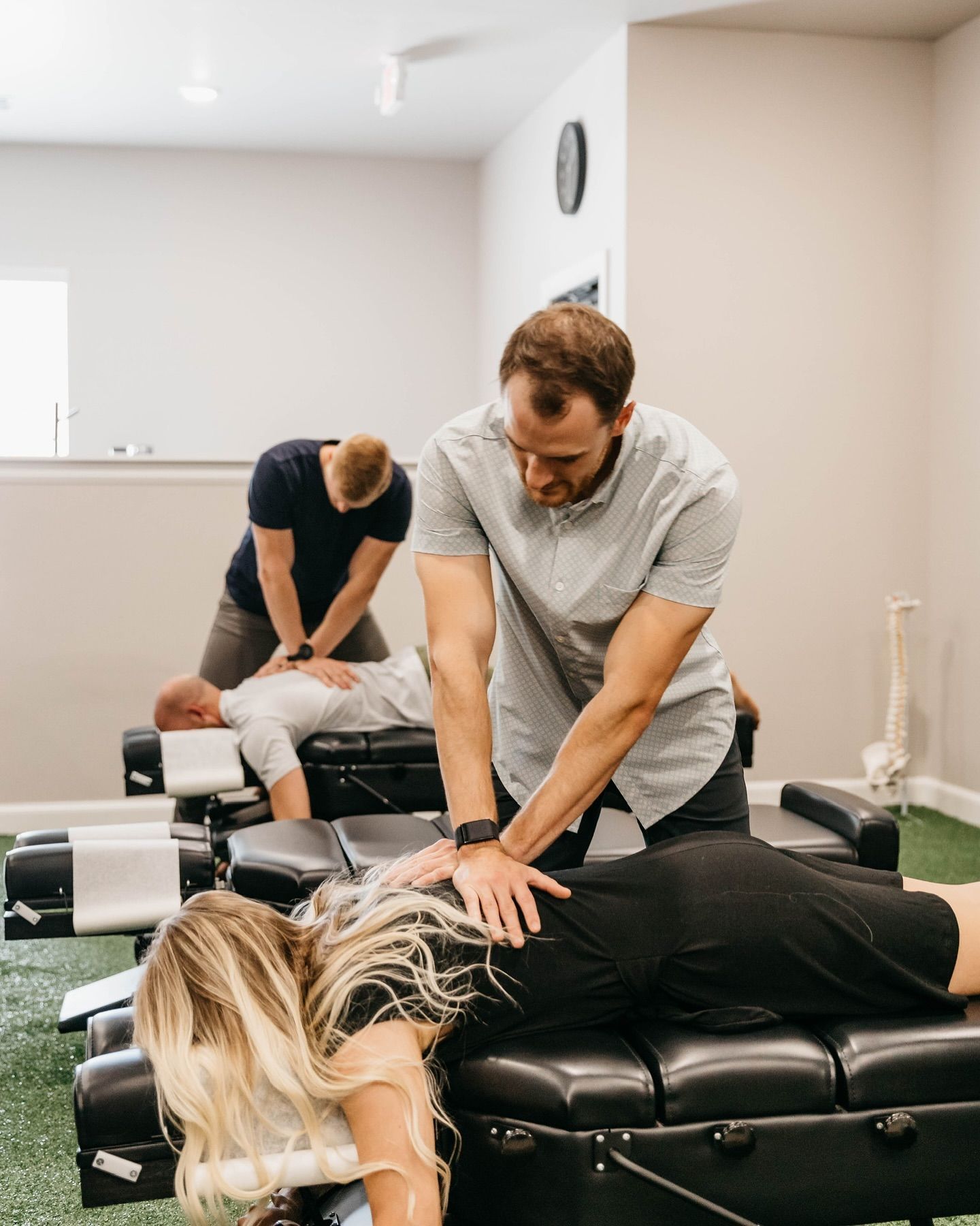 Massage therapists working on a client in a clinic room with treatment tables and wall clock