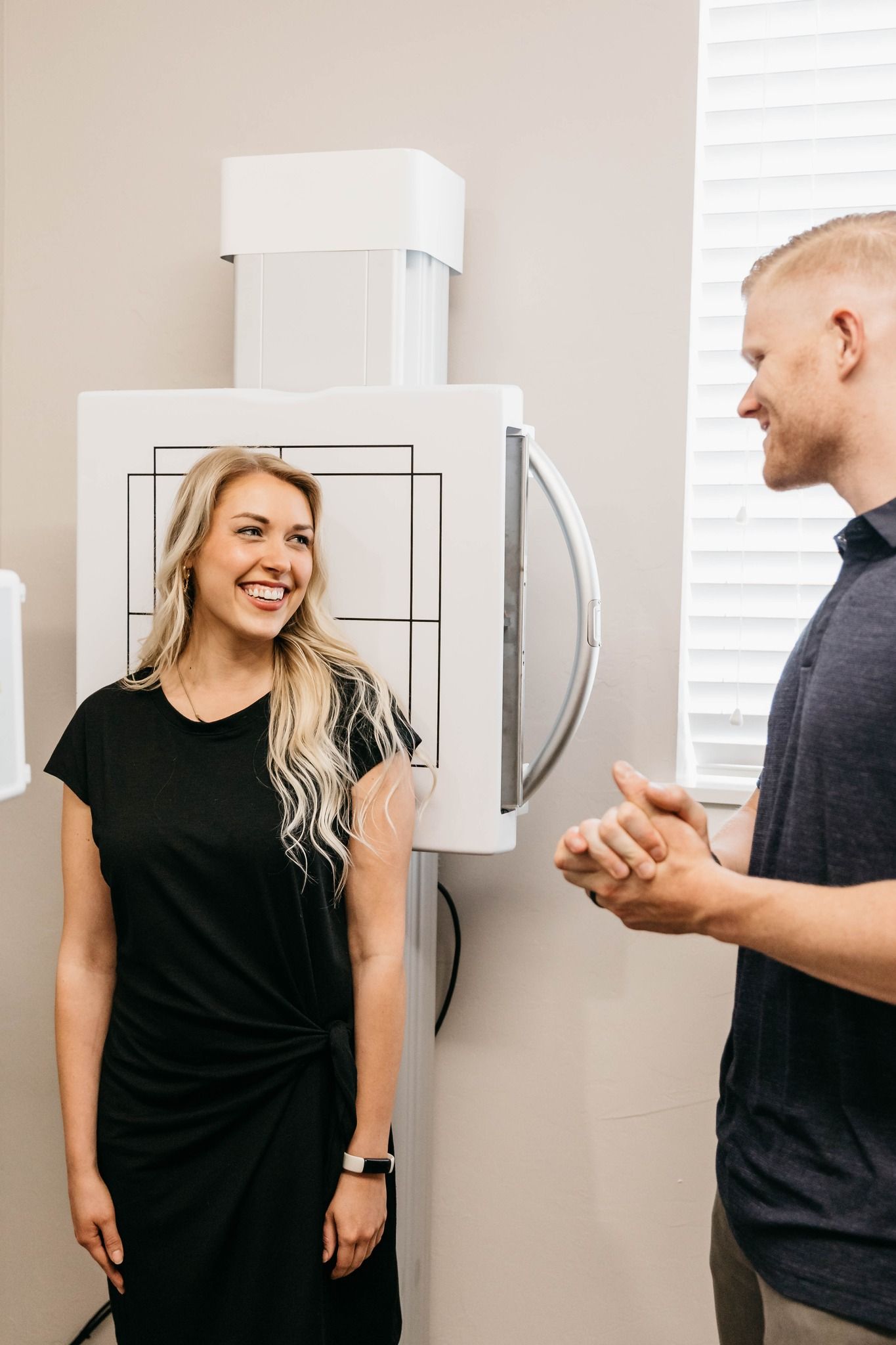 Woman smiling beside X-ray machine while man speaks with her in a medical office