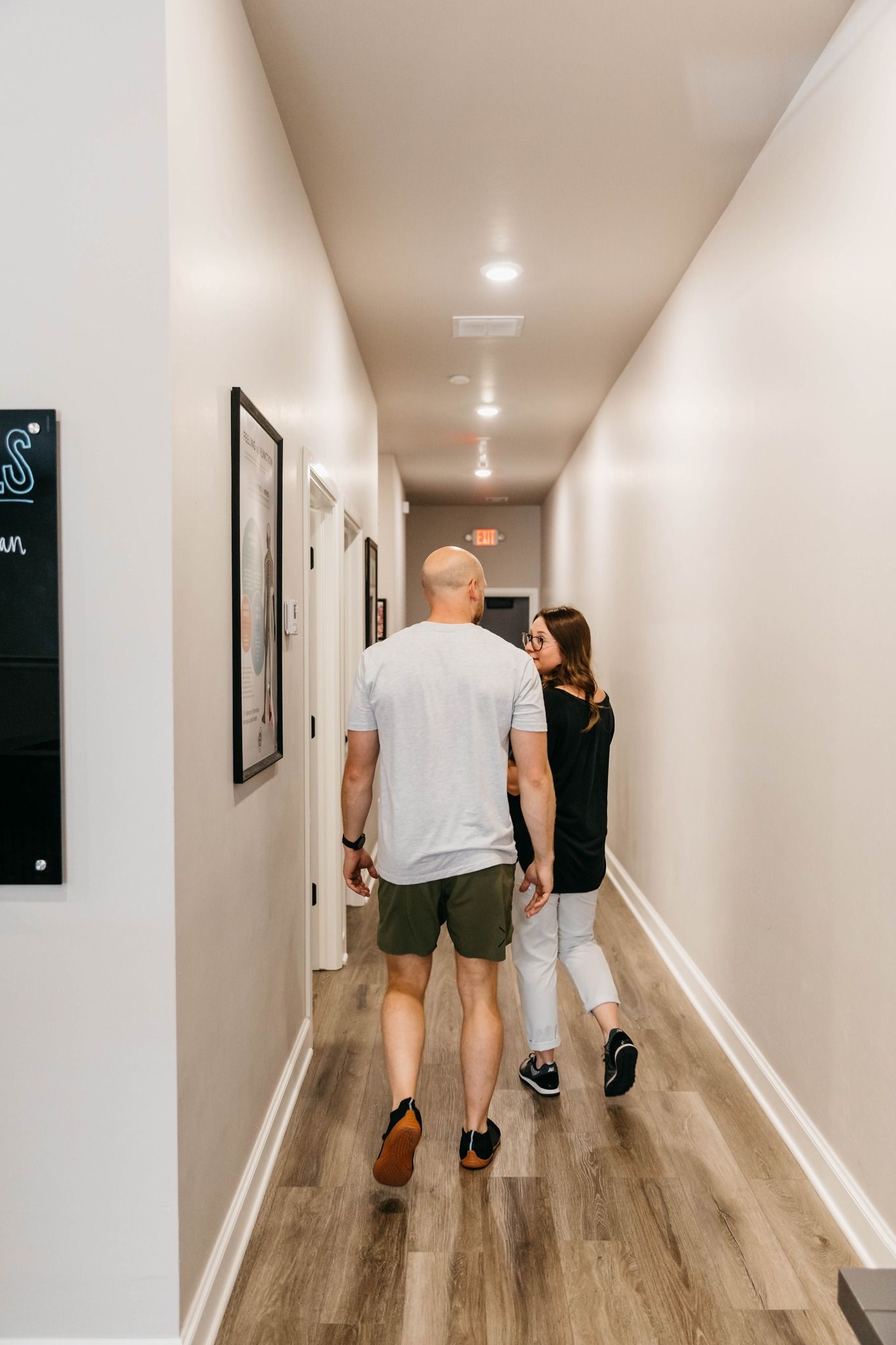Two people walking down a narrow hallway with white walls and wood flooring