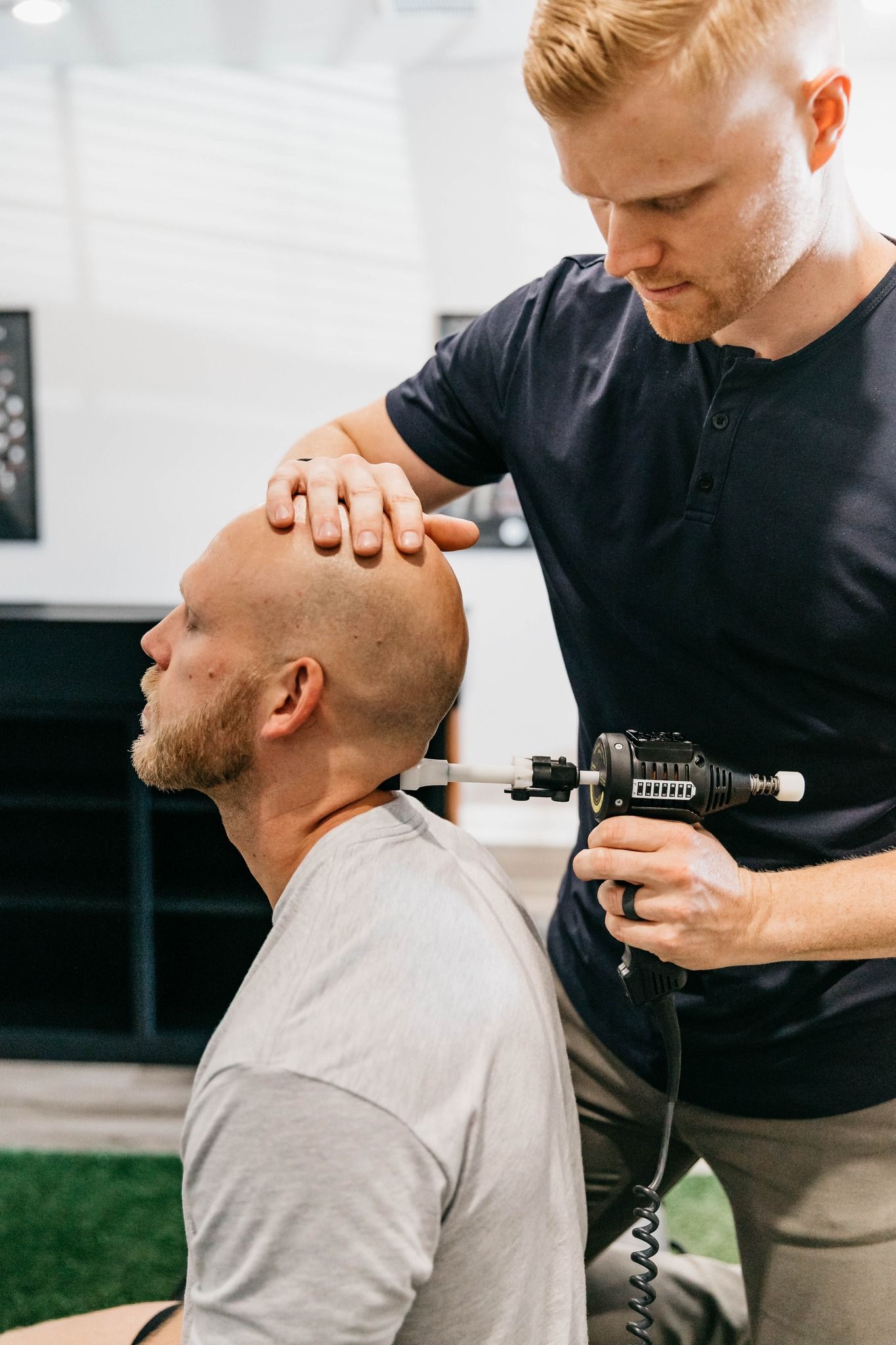 Trainer adjusting a bald client's neck with a handheld device in a gym setting