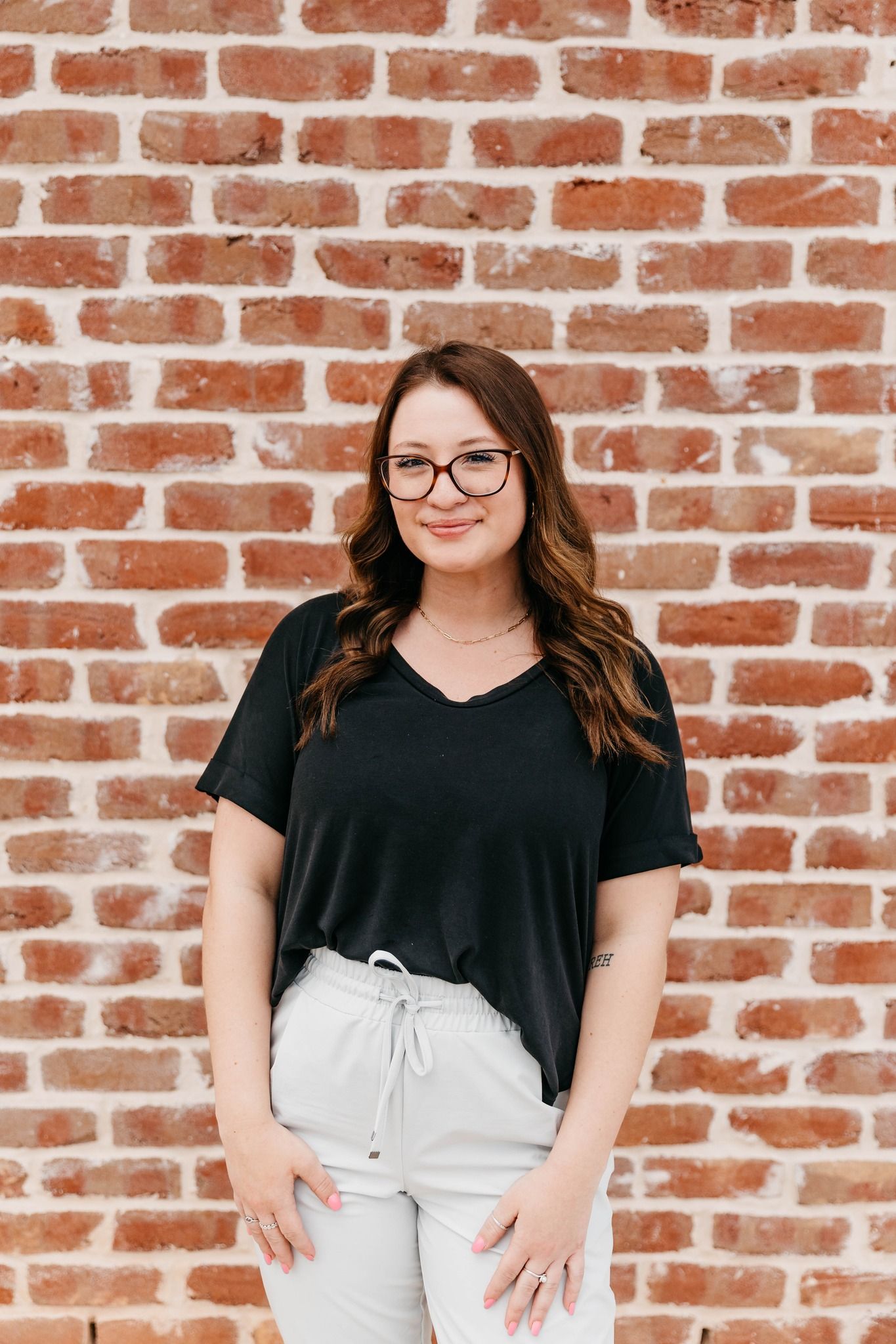 Woman standing against a brick wall, wearing a black top and light pants, smiling at the camera.