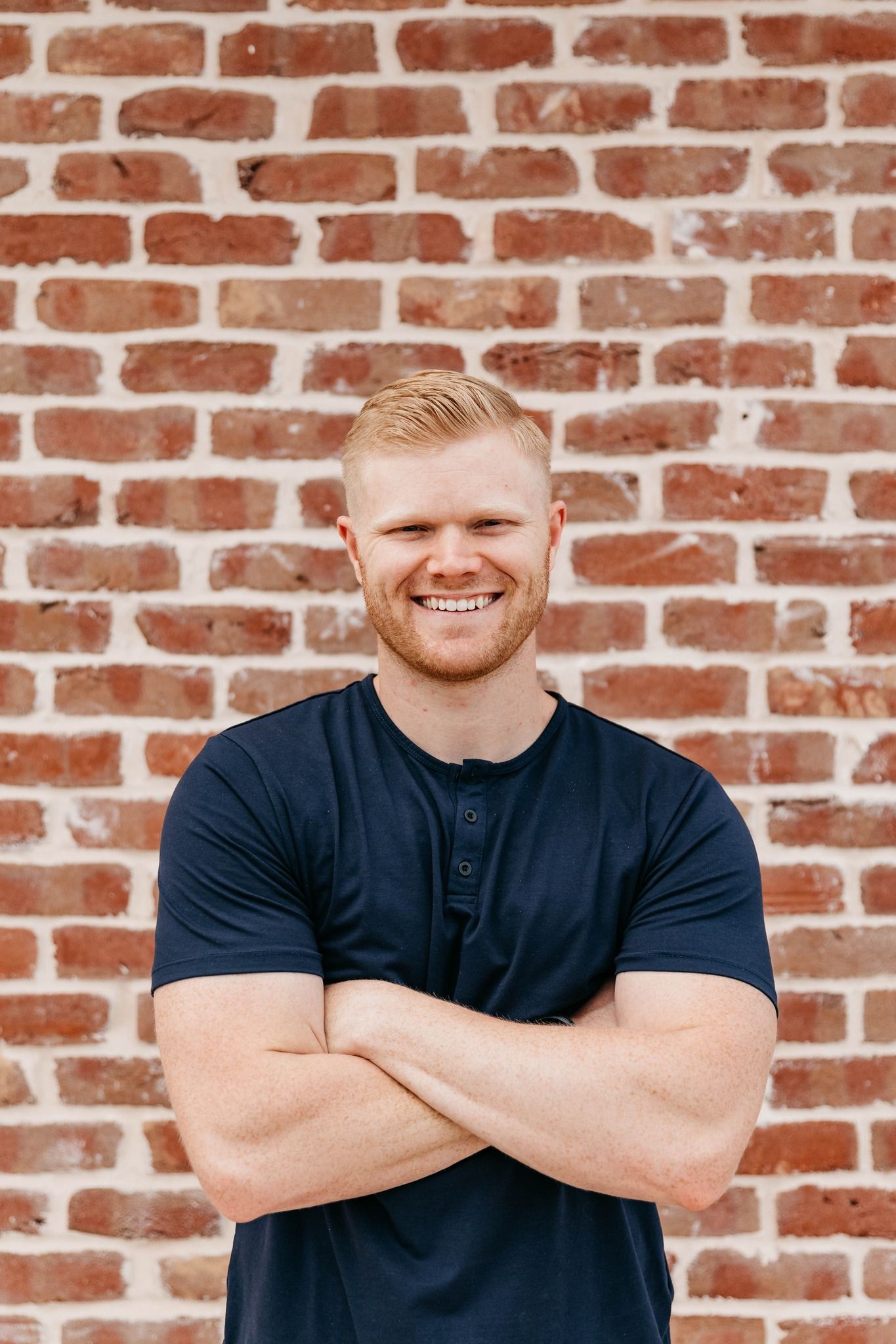 Smiling man in a dark shirt standing with arms crossed against a red brick wall