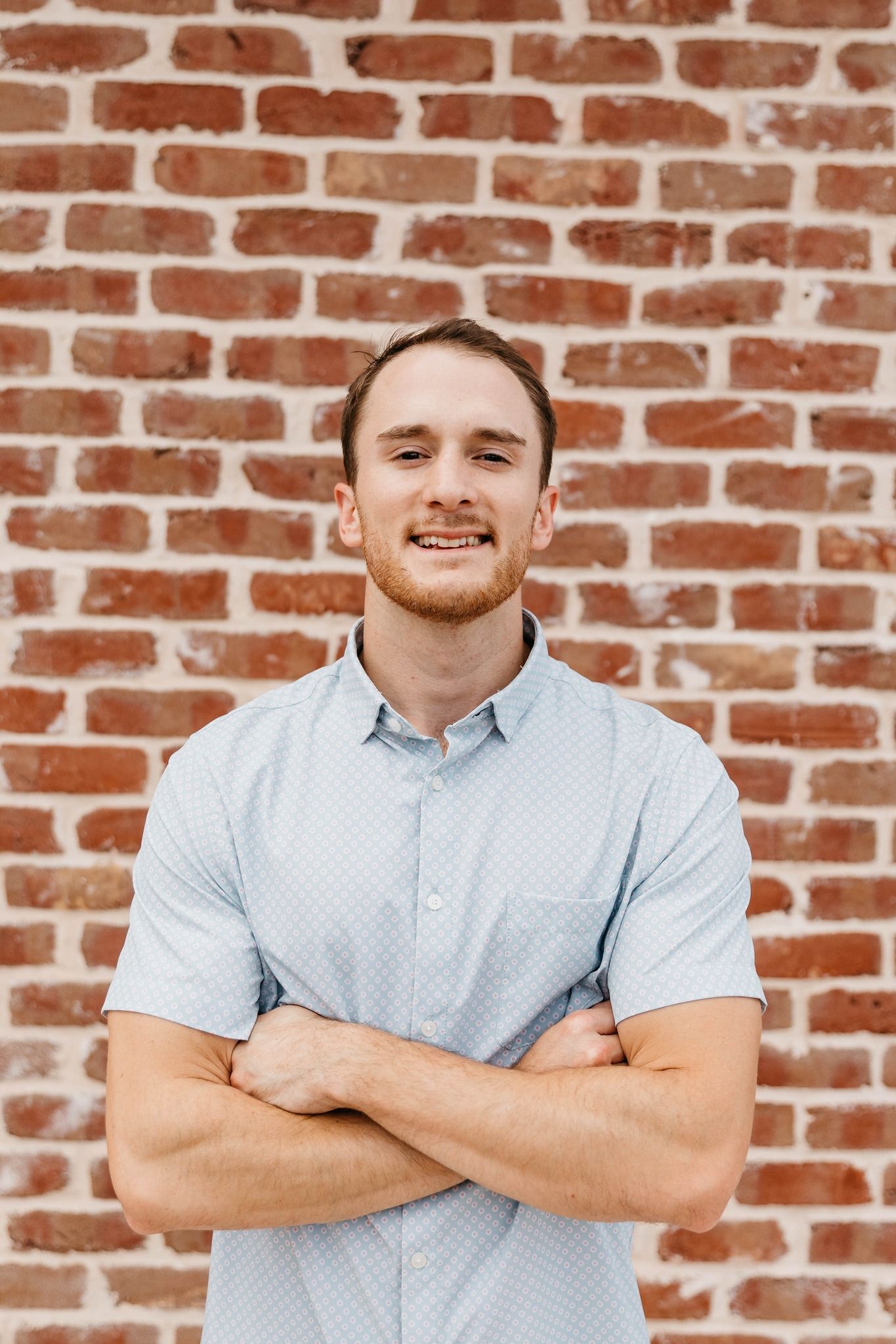 Smiling person in a light blue shirt, arms crossed, standing against a brick wall.