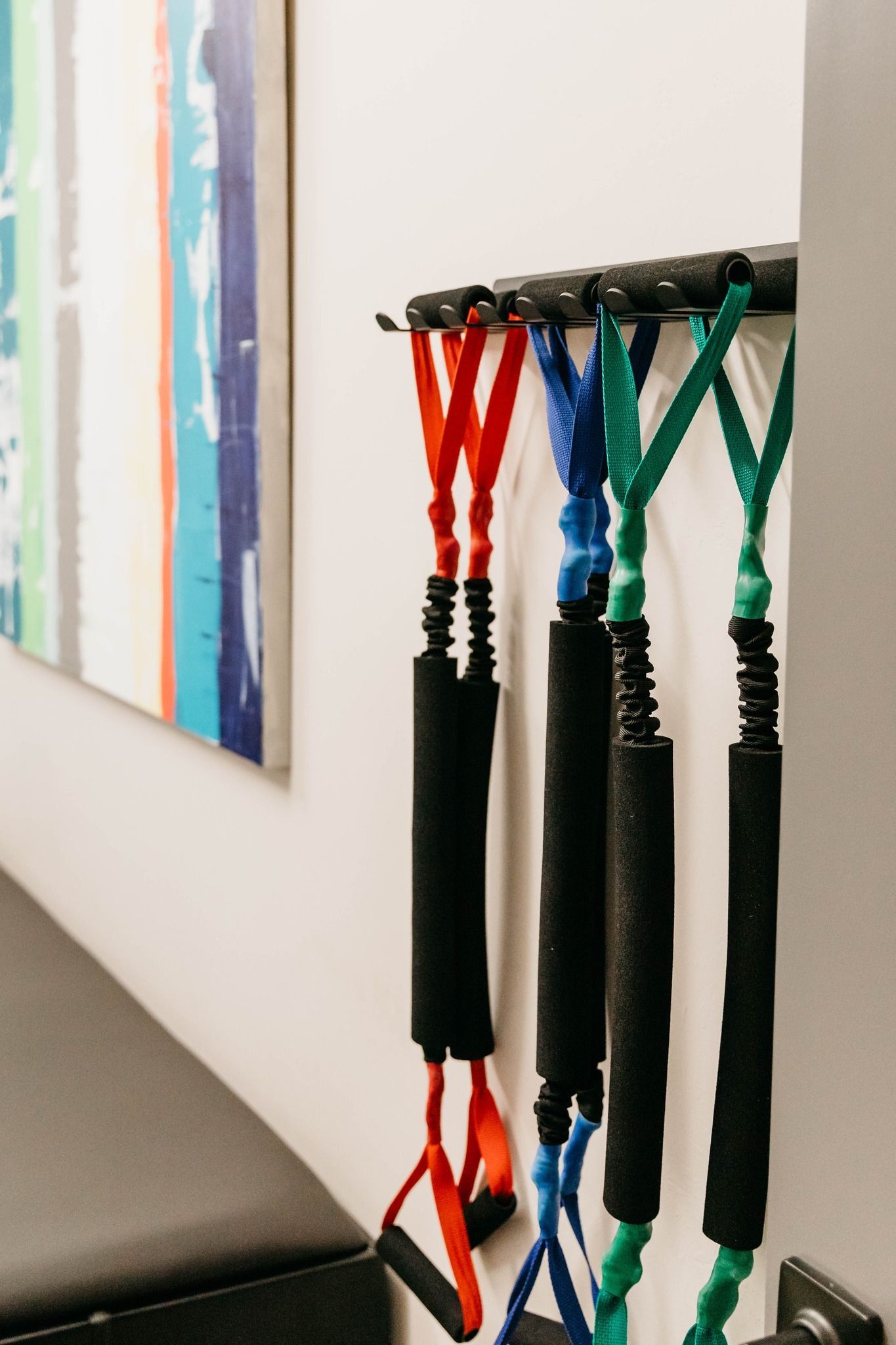 Colorful braided exercise handles hanging on a wall-mounted rack beside framed artwork.