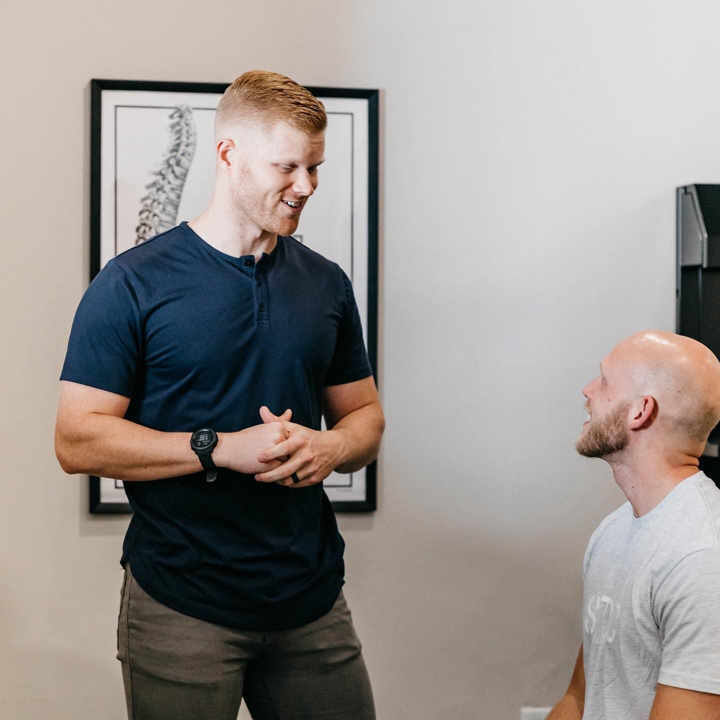 Two men talking in a room, one standing and smiling, the other seated and looking up.