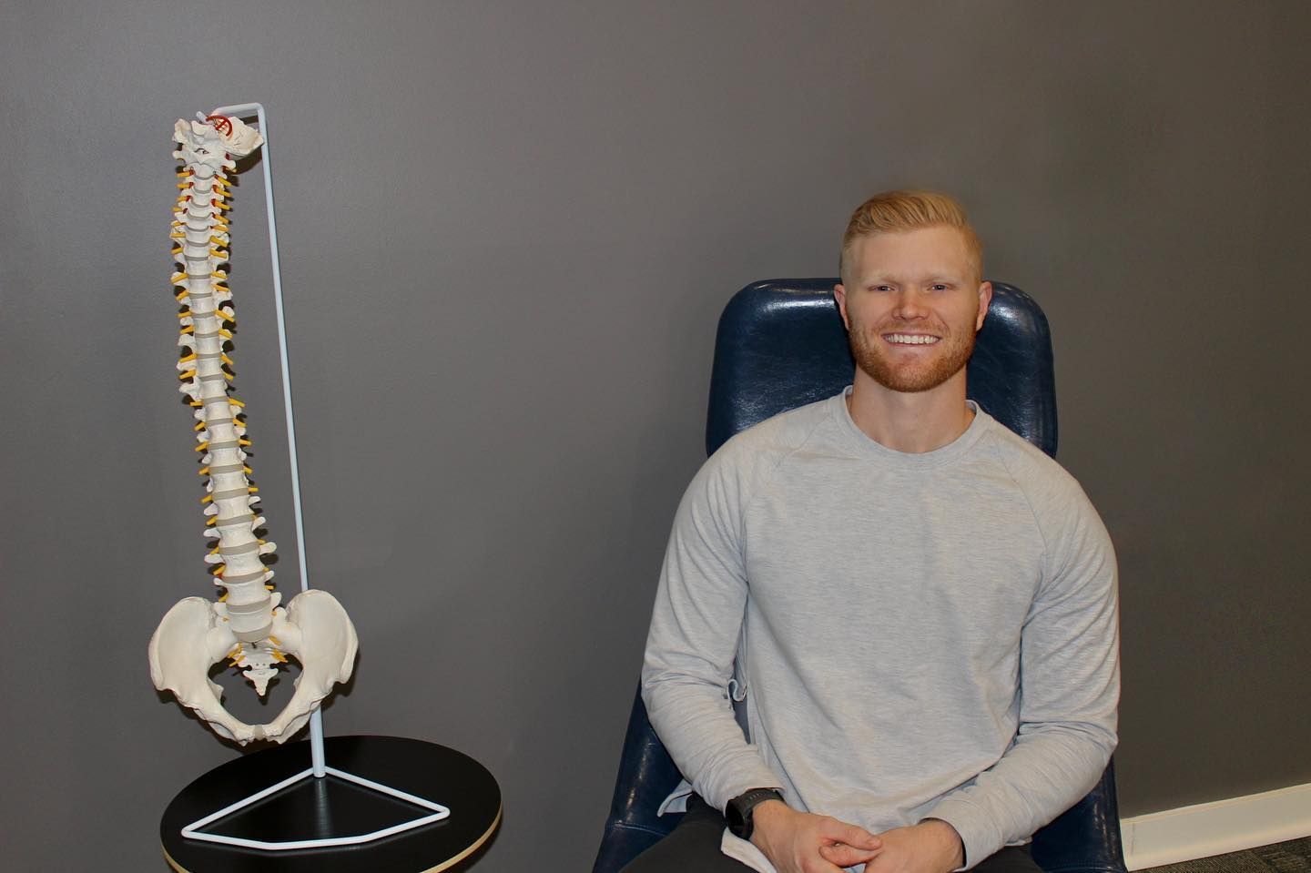 Man sitting in a chair beside a model human spine on a stand in a plain room