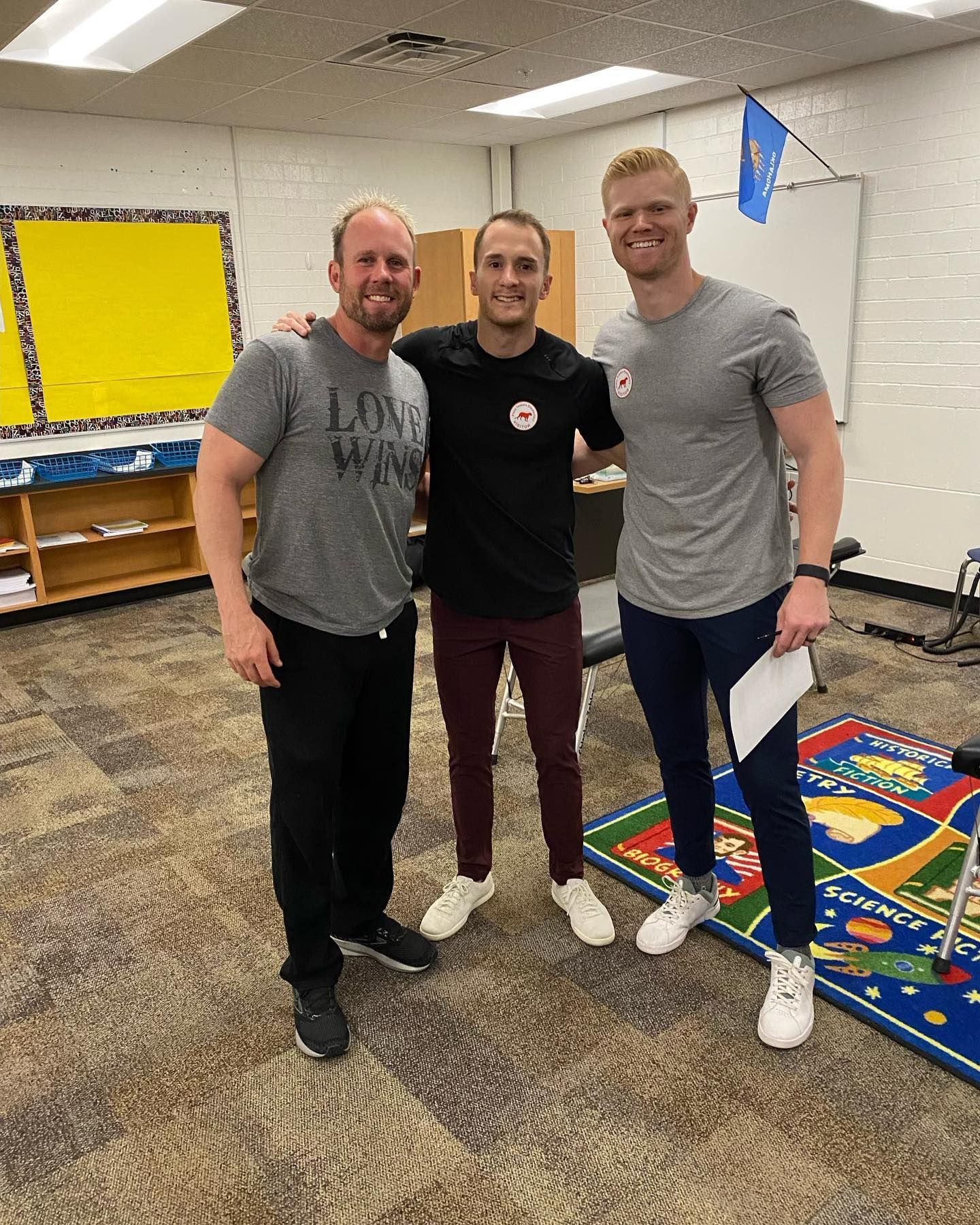 Three men standing together in a classroom beside a colorful rug and bulletin boards.