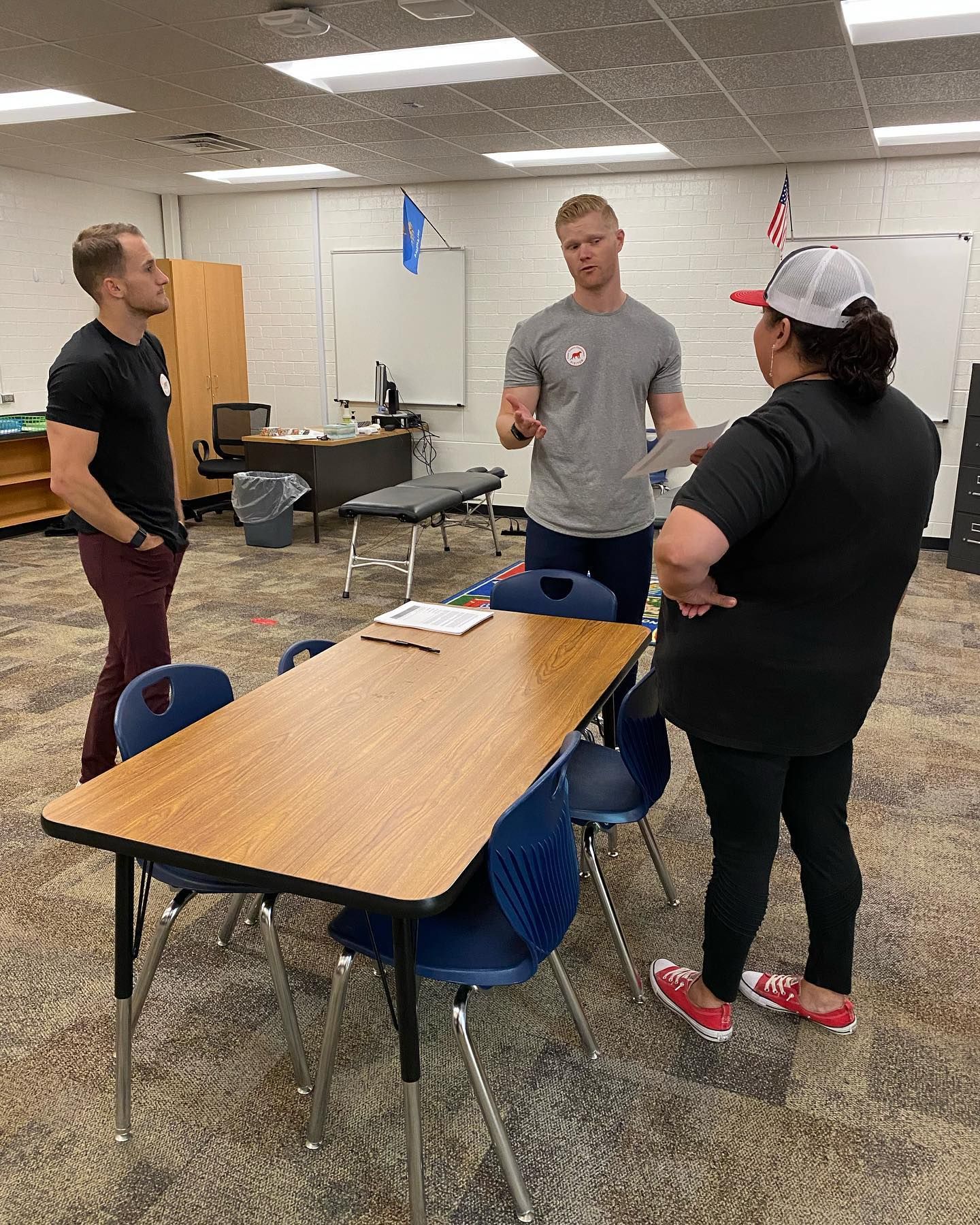 Three people stand around a table in a classroom, talking during a casual meeting.