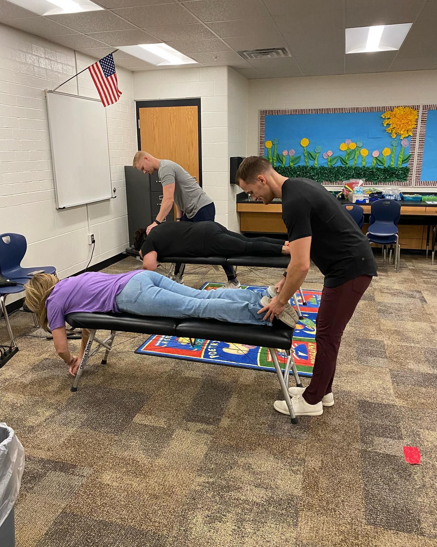 Two people support a person in a purple shirt doing a horizontal plank in a classroom