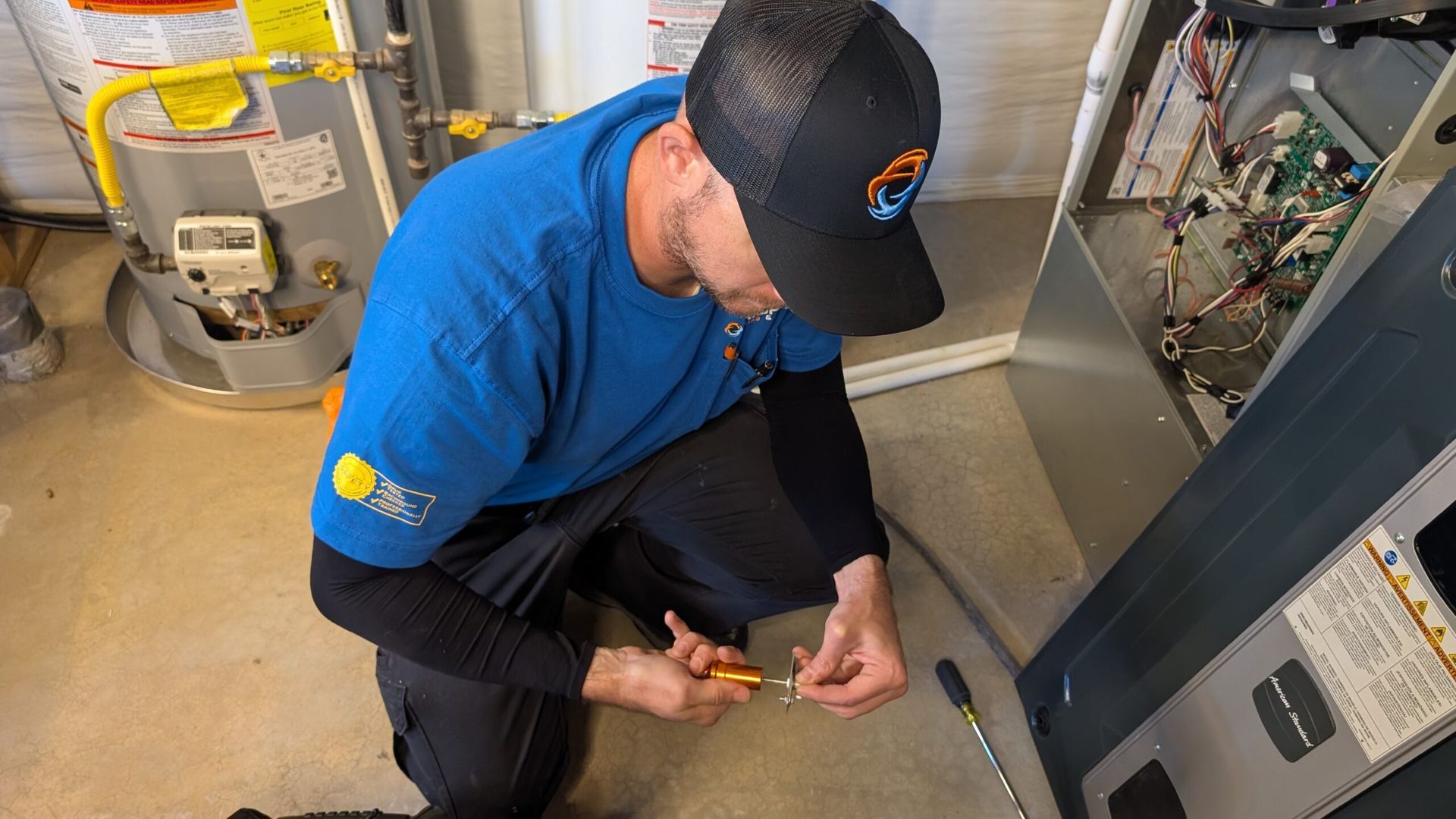 Worker in blue shirt kneels by electrical panel, using a tool on wiring in a utility room.