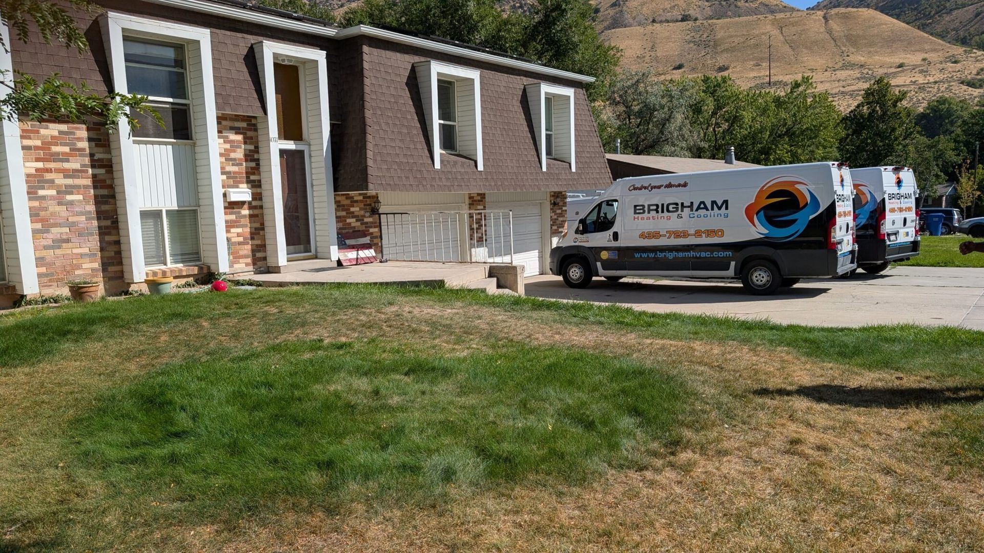 House with landscaping truck parked in driveway, beside a grassy yard and rocky hills.