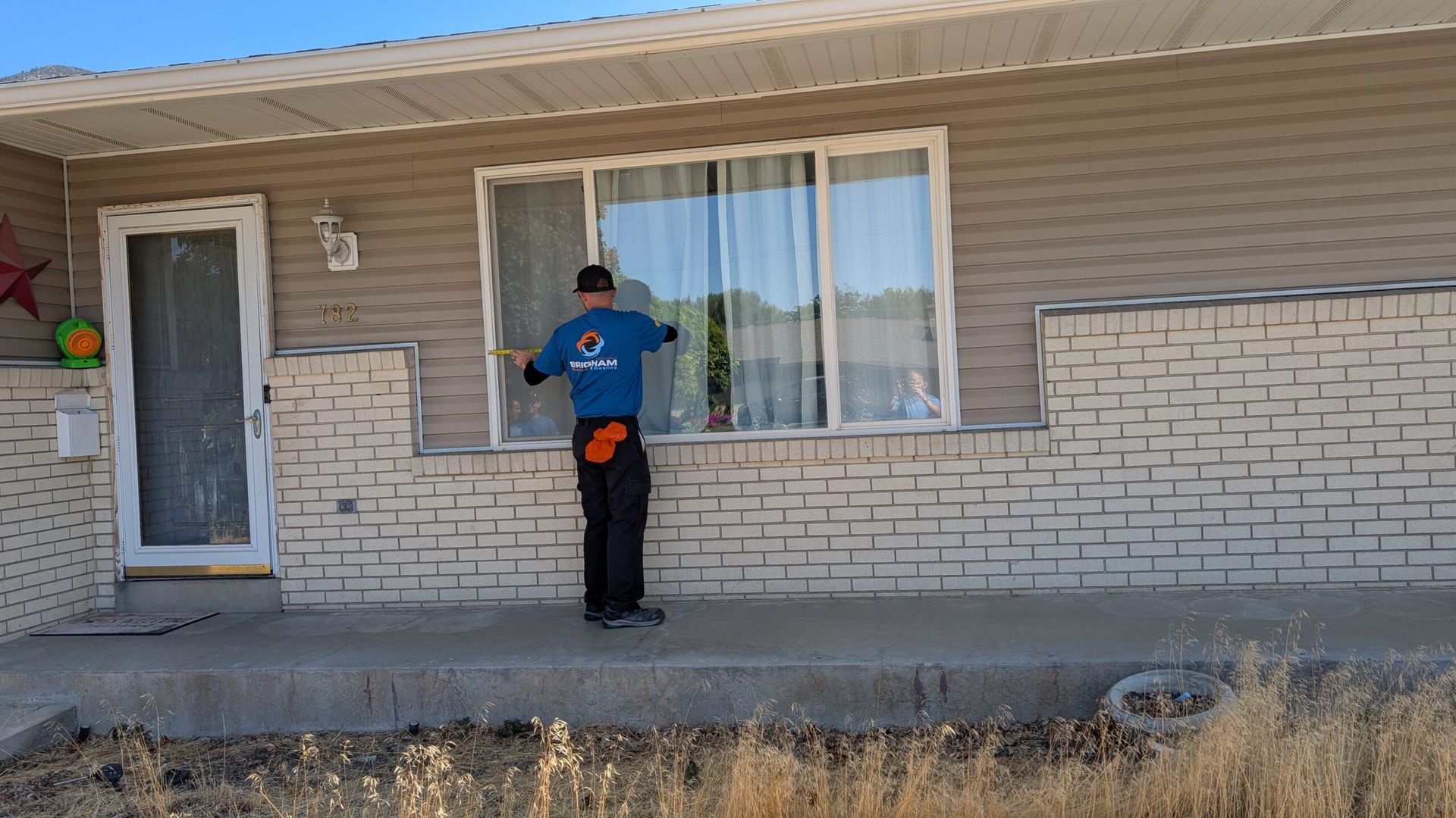 Person standing on a porch next to a window on a beige house, wearing a blue and orange shirt.
