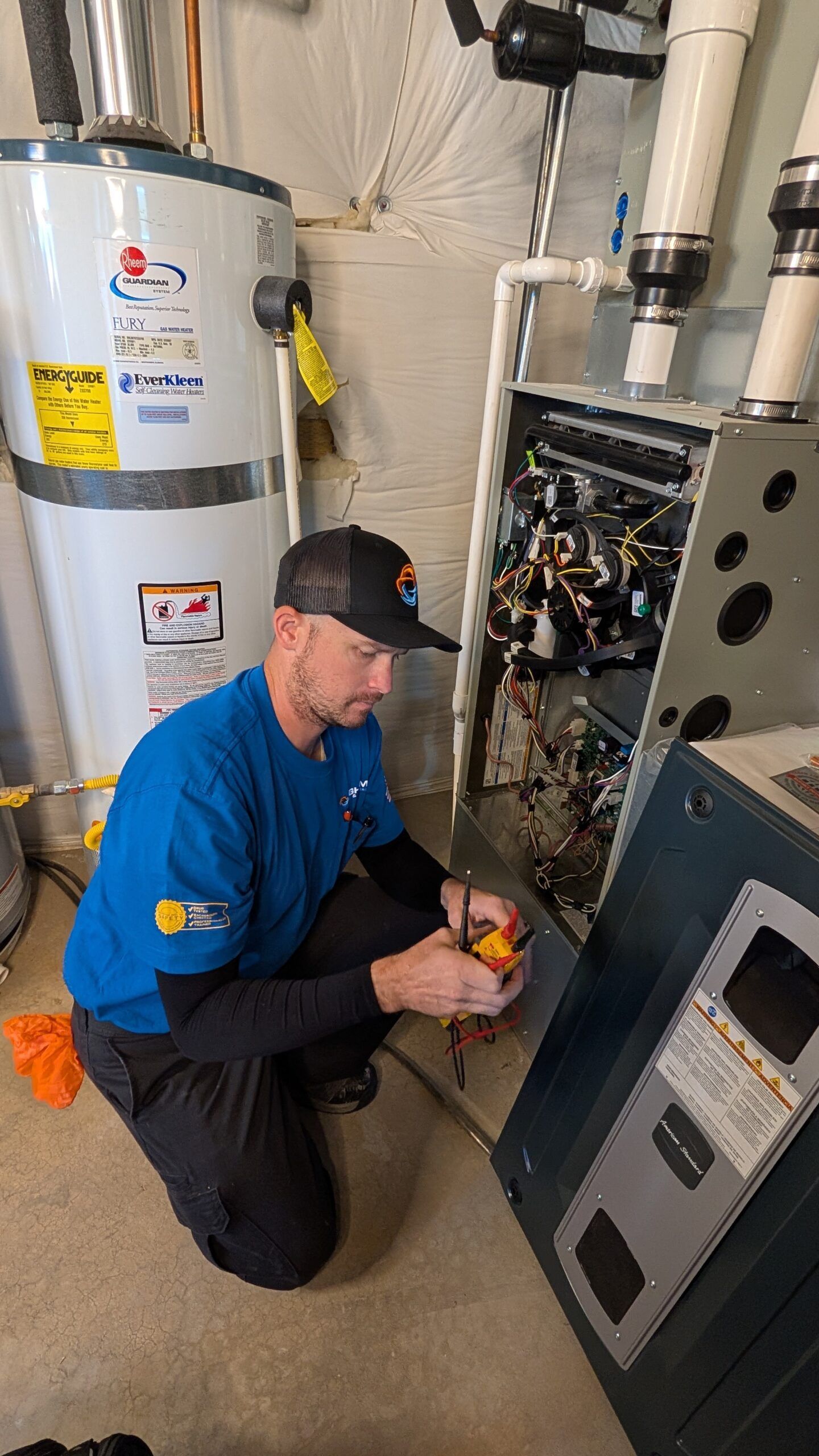 Technician kneeling beside a water heater, inspecting an open HVAC unit with tools in a utility room
