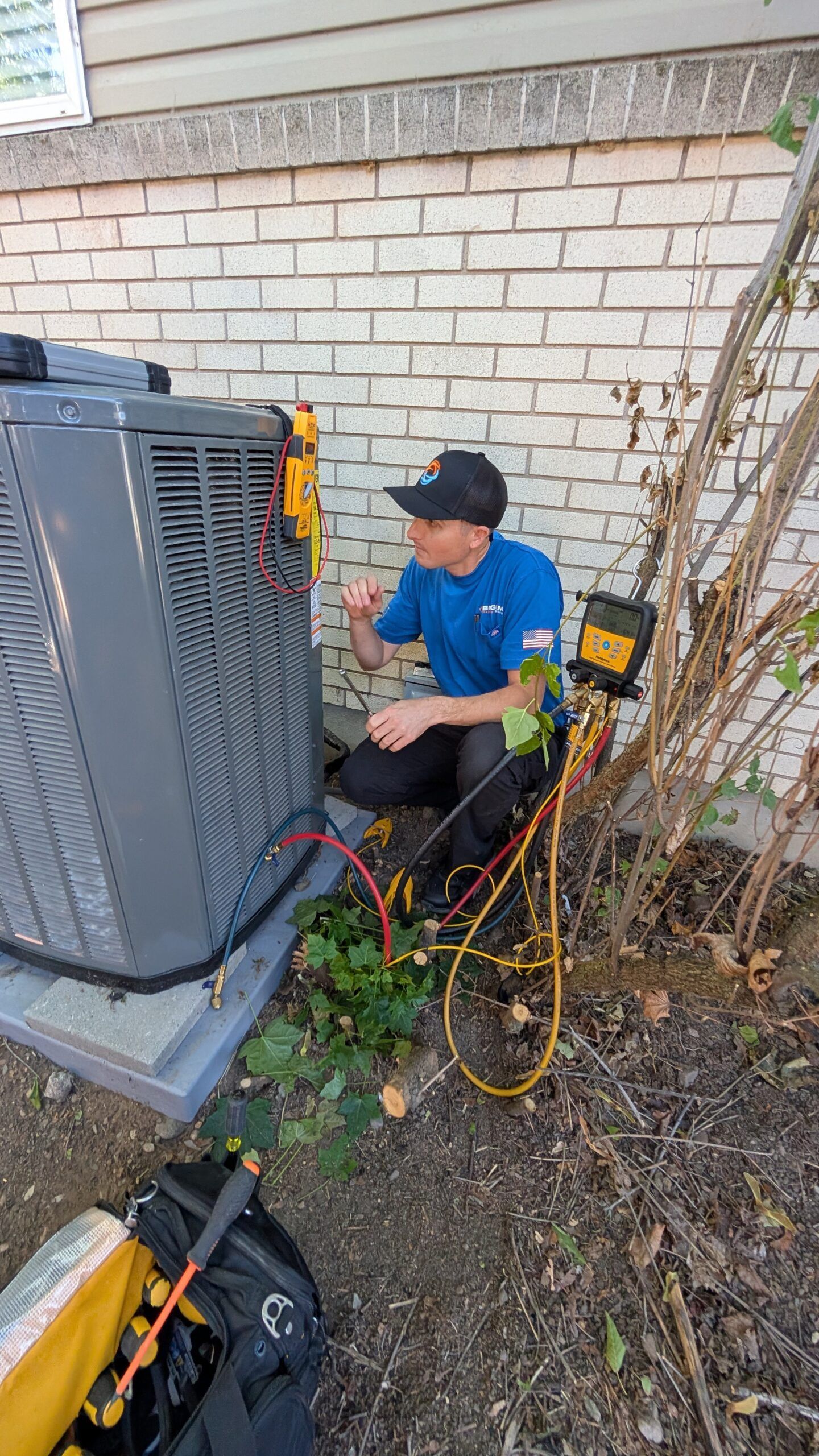Technician kneeling beside an outdoor HVAC unit, checking equipment and tangled cables against a brick wall.