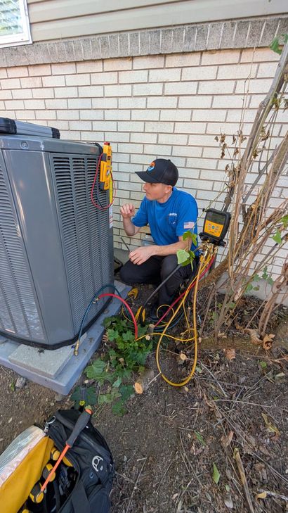 Technician kneeling beside an outdoor HVAC unit, checking equipment and tangled cables against a brick wall.