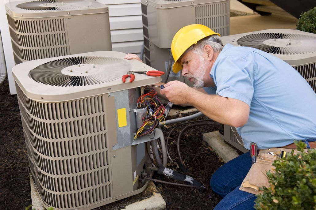 Technician in a yellow hard hat repairs an outdoor air conditioner unit beside several others.