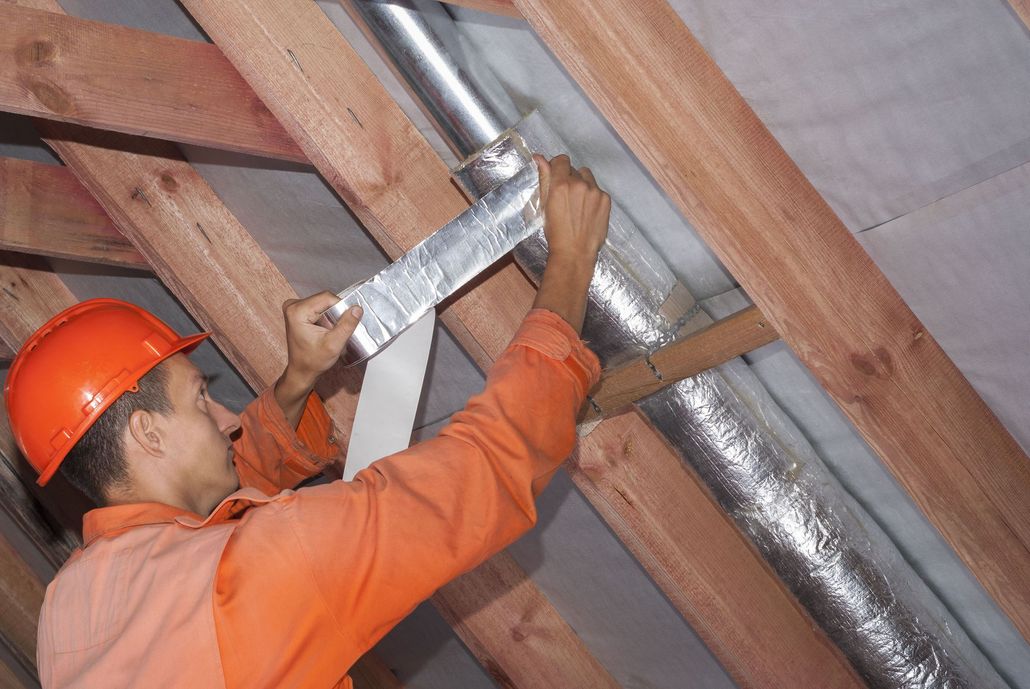 Worker in orange hard hat installing shiny ducting in attic framing