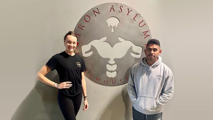 A man and a woman are standing in front of a logo for Iron Asylum Gym