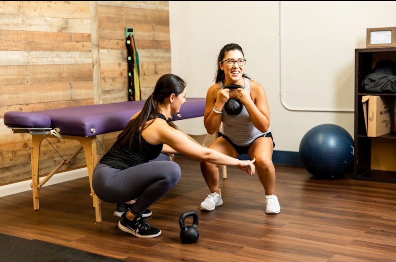A woman is squatting next to another woman in a gym while holding a kettlebell