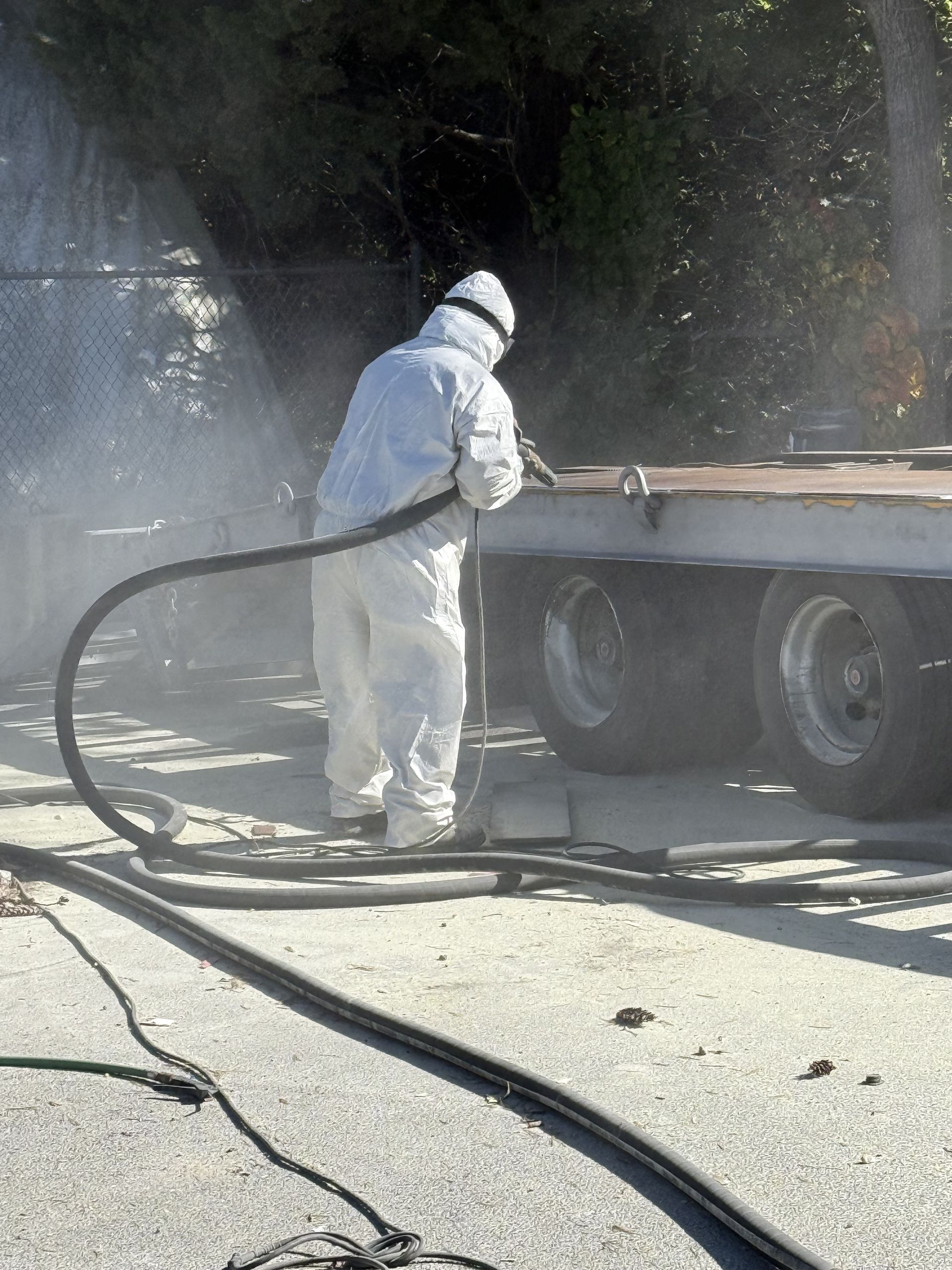 Person in white protective suit sandblasting a trailer outdoors, visible spray.