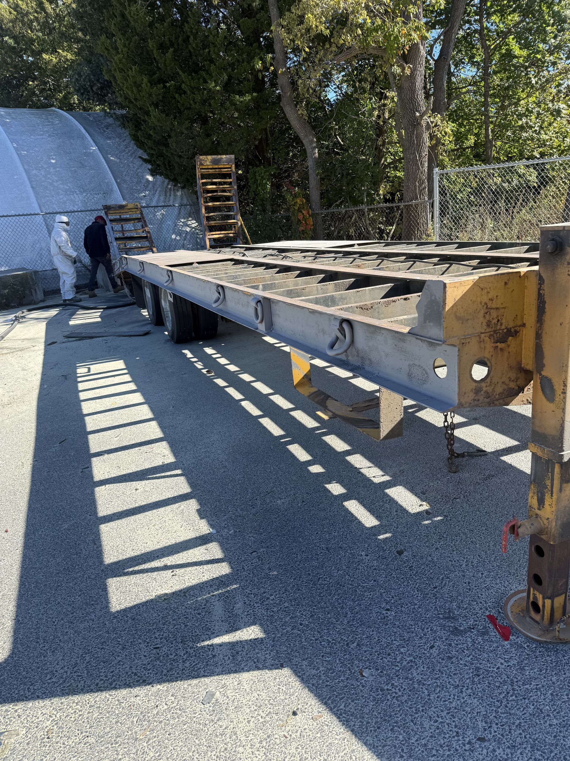 Flatbed trailer on gravel, with a person in protective suit nearby. Trees in the background.