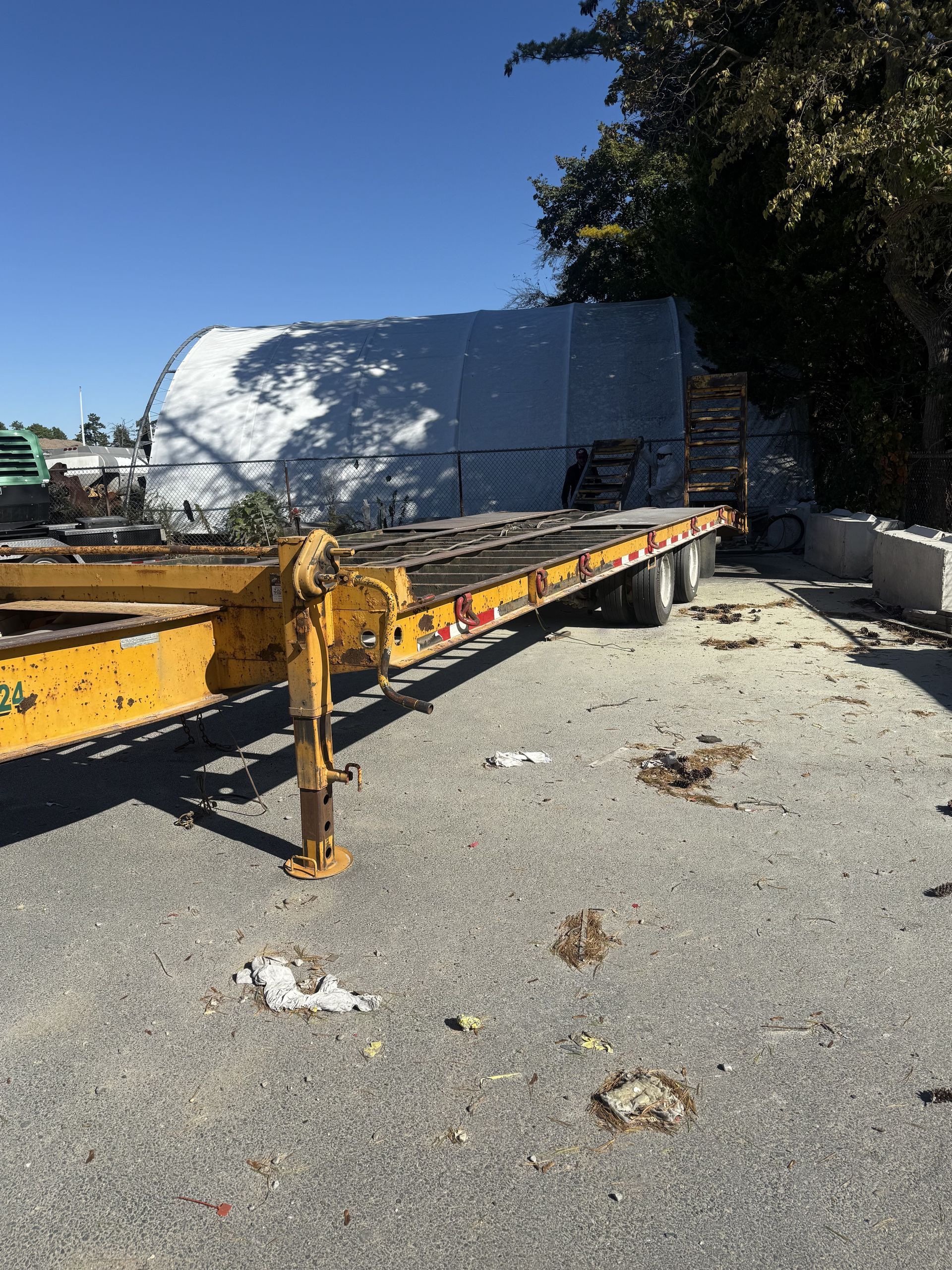 Yellow flatbed trailer on a gravel lot under a blue sky, with a white structure in the background.