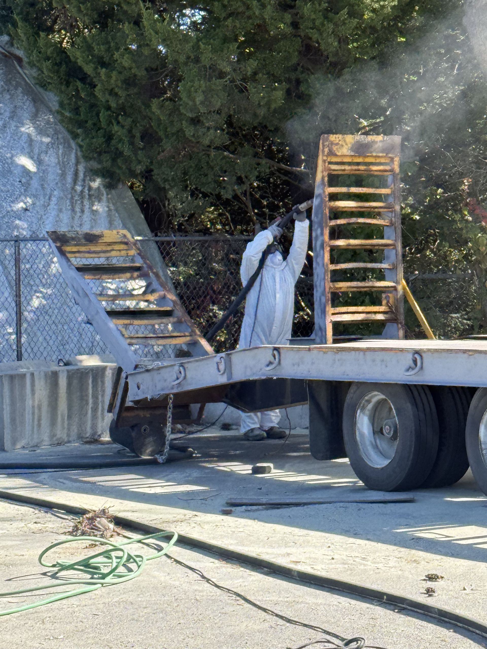 Person in protective suit sandblasting metal structure on a flatbed trailer outdoors. Spraying dust.