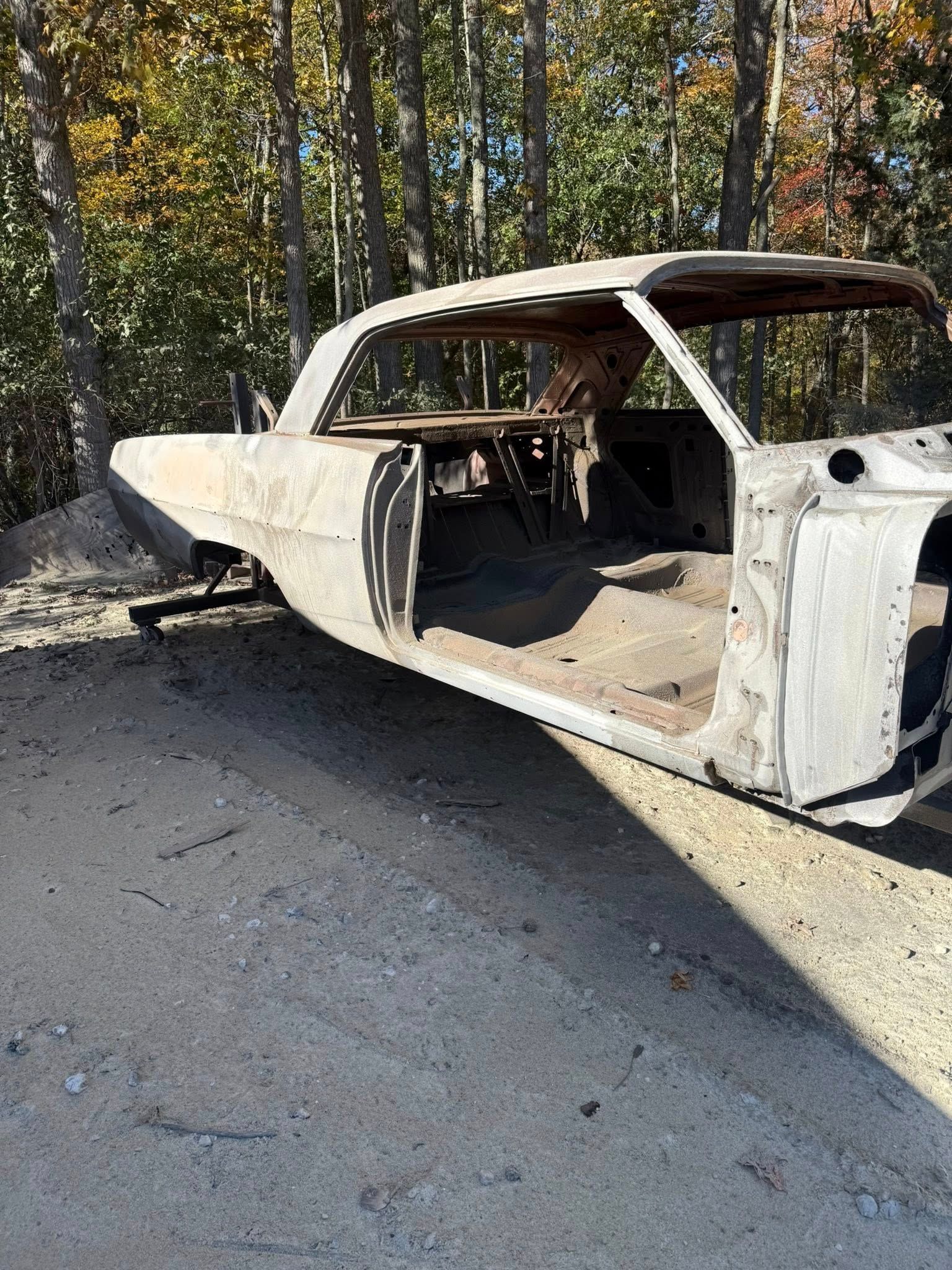Bare metal car body on a gravel surface, wooded background.