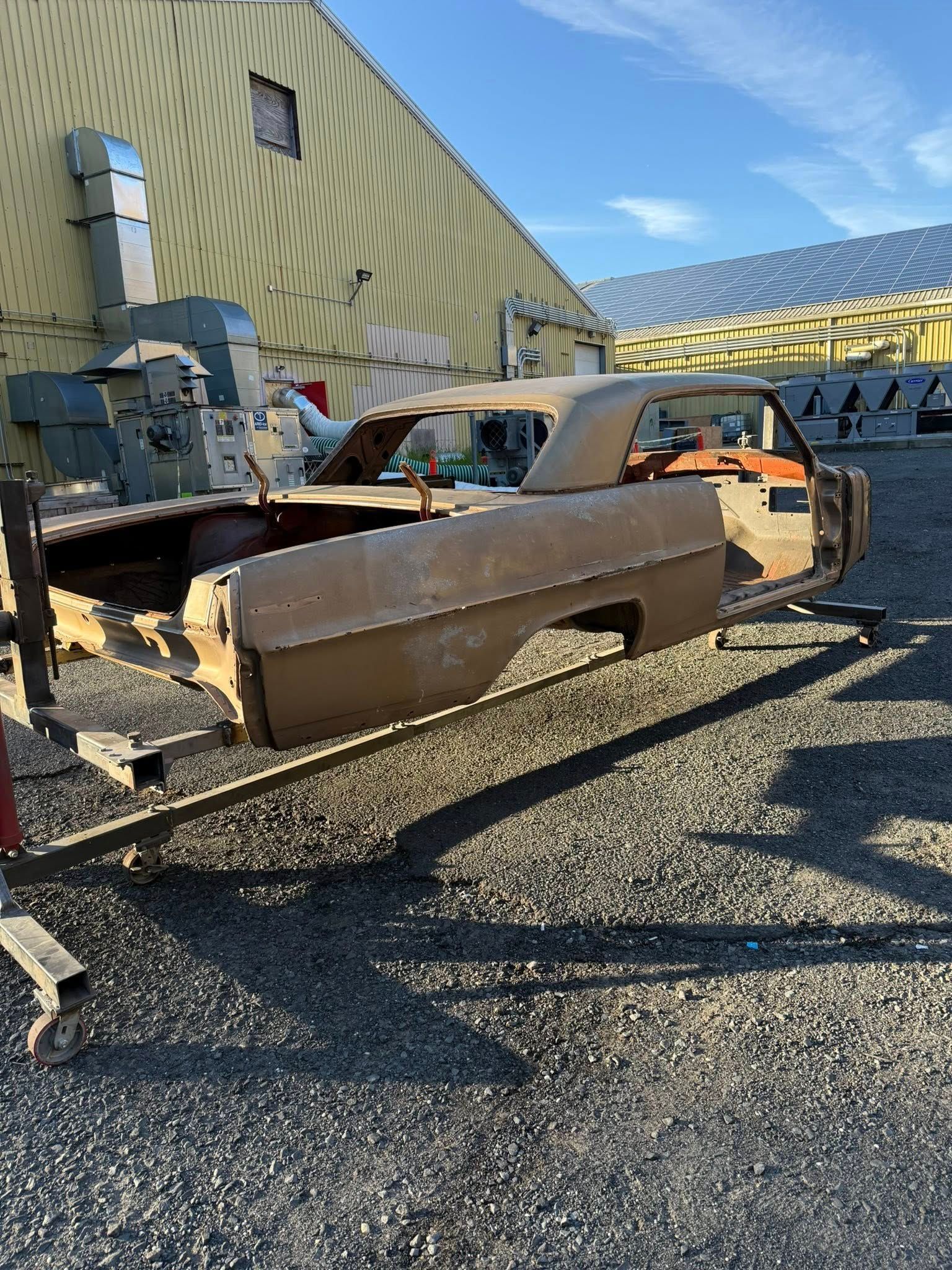 Brown car body on a cart, partially dismantled, outdoors near a warehouse on a gravel lot.