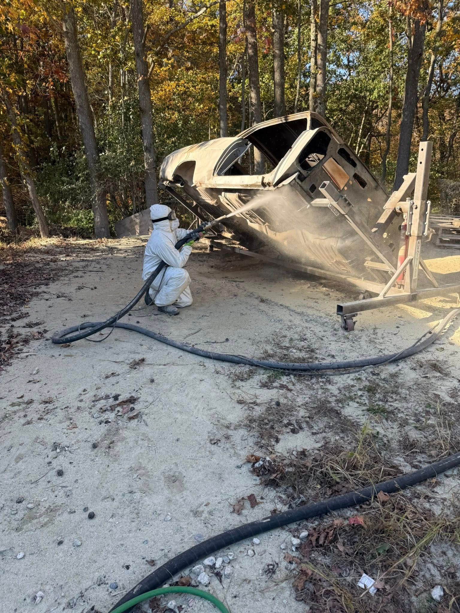 Person in protective suit sandblasting vehicle frame outdoors.