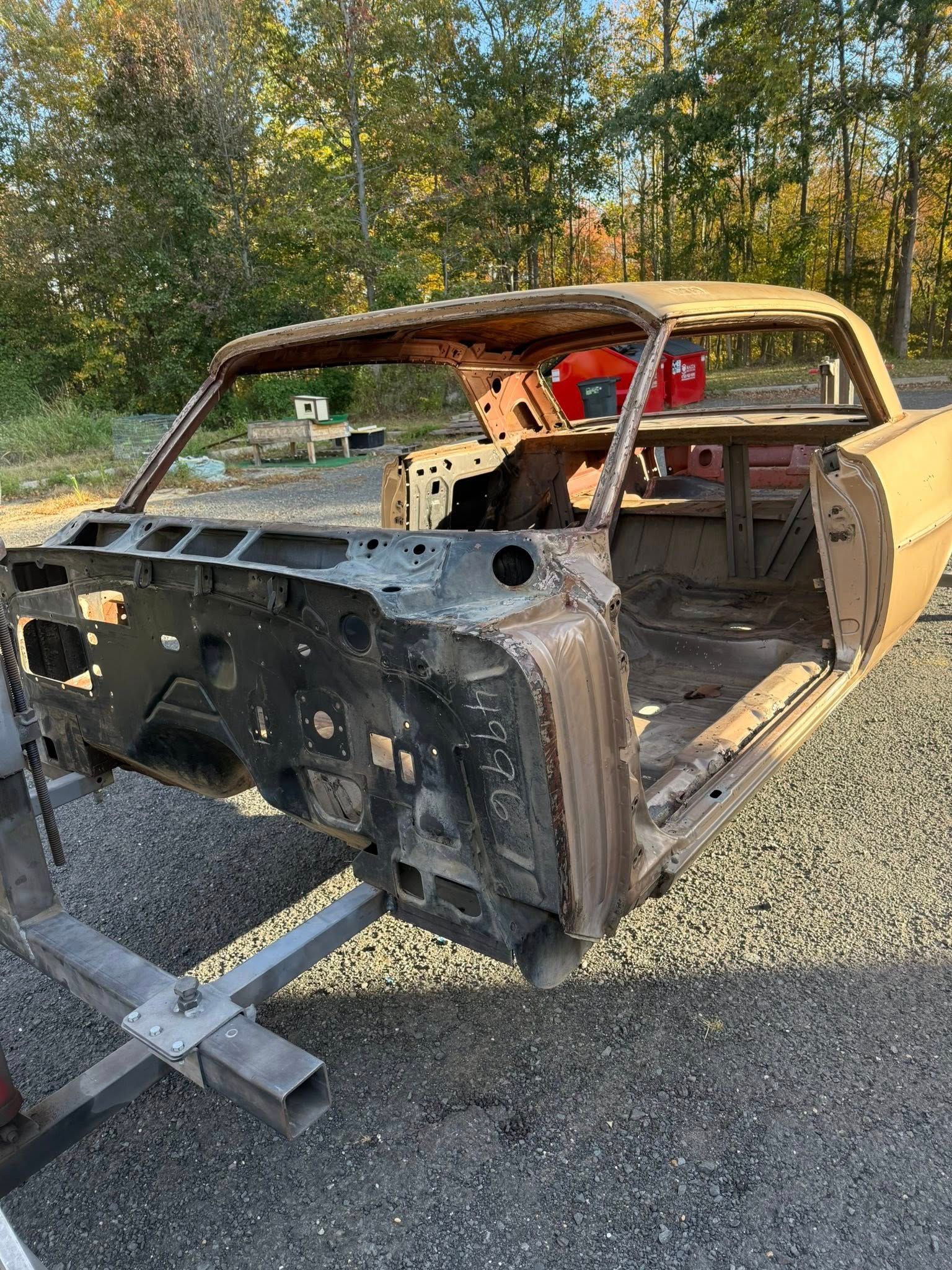 Bare frame of an old gold-colored car body, on a lift in an outdoor setting.