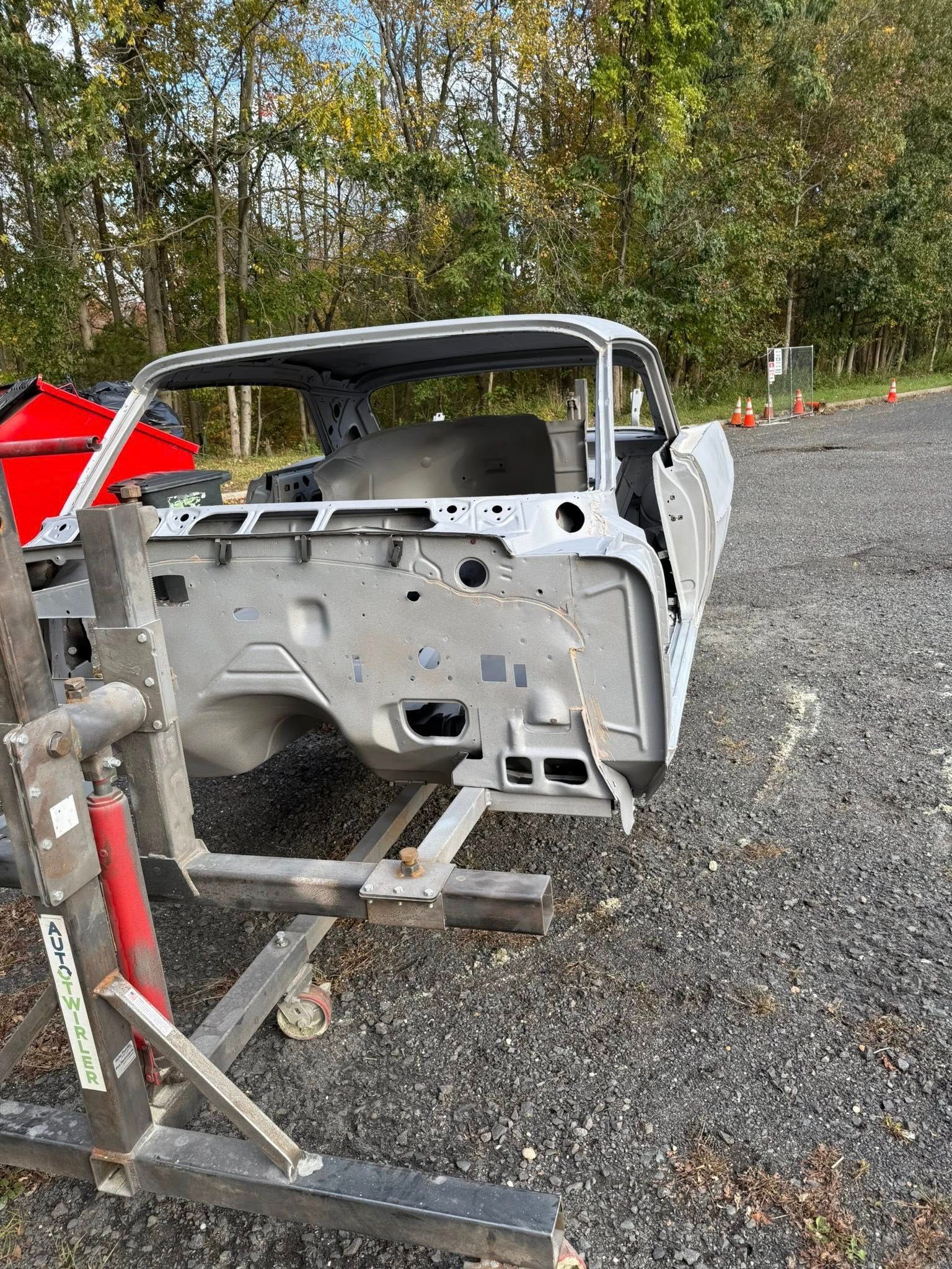 Bare car chassis on a lift, outdoors. Gray metal, trees in background.