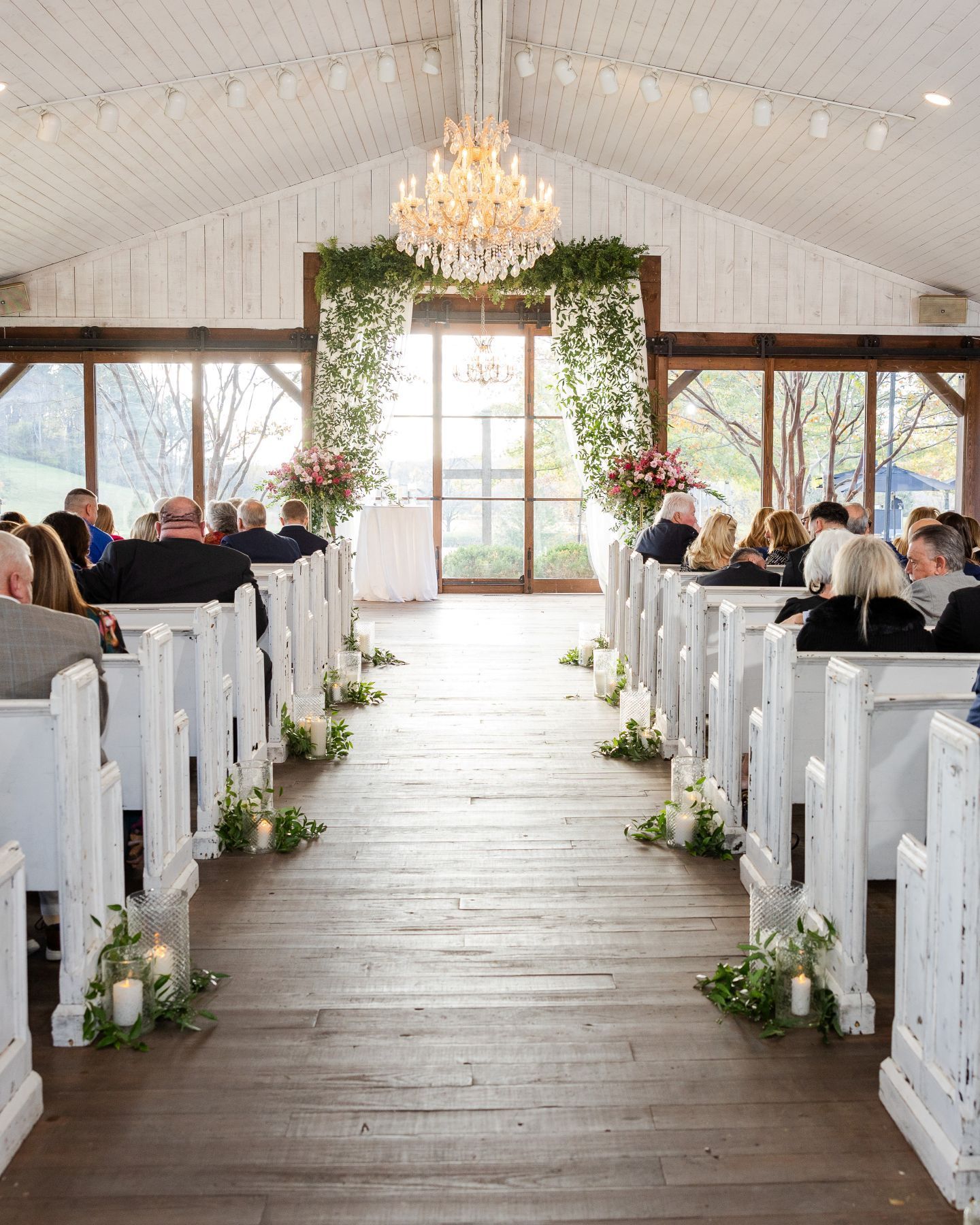 Wedding ceremony in a bright, rustic venue with white pews, floral arrangements, and a chandelier. Guests are seated.