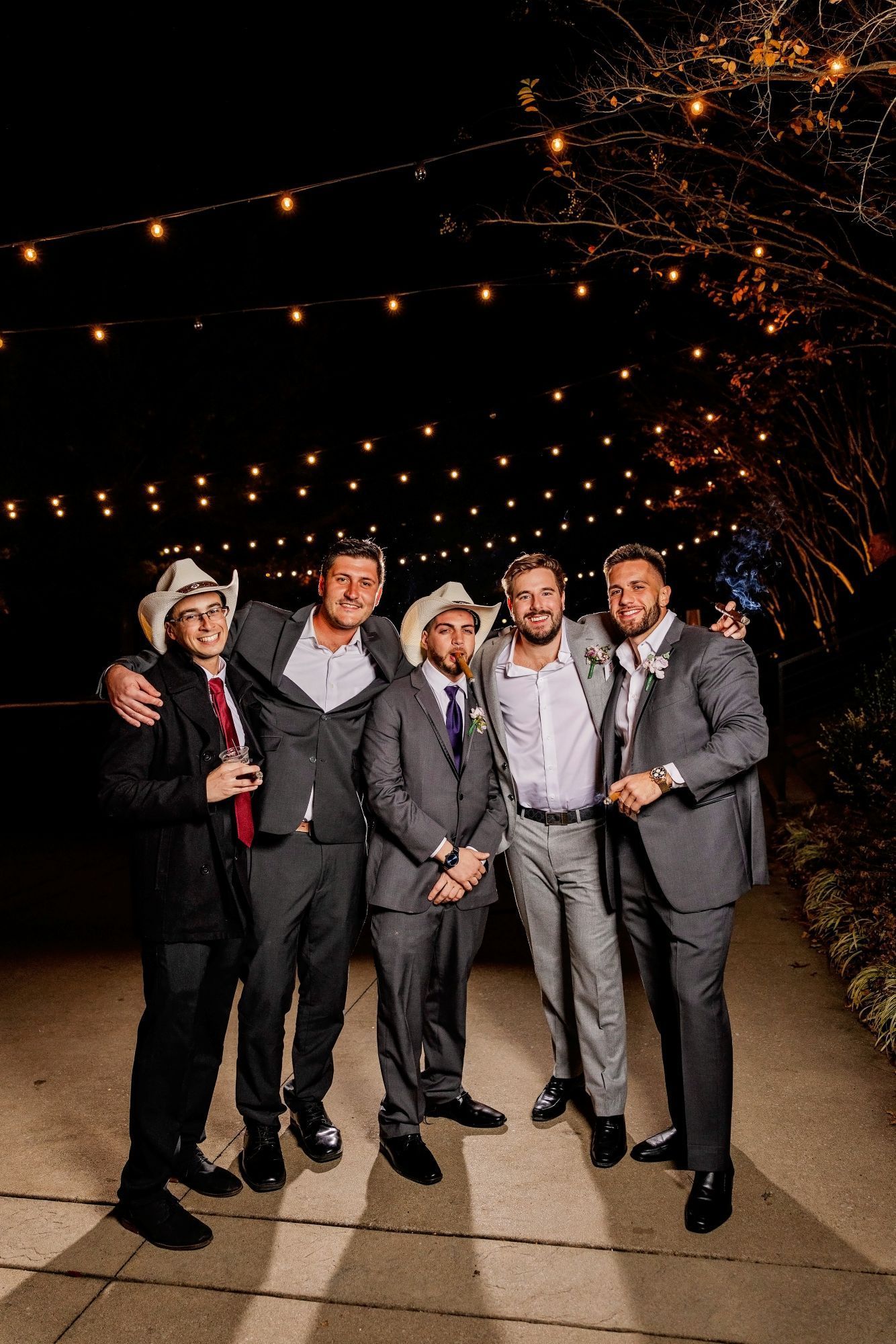 Five men in suits and cowboy hats pose on a path at night, lit by string lights. Some have arms around each other, smiling.